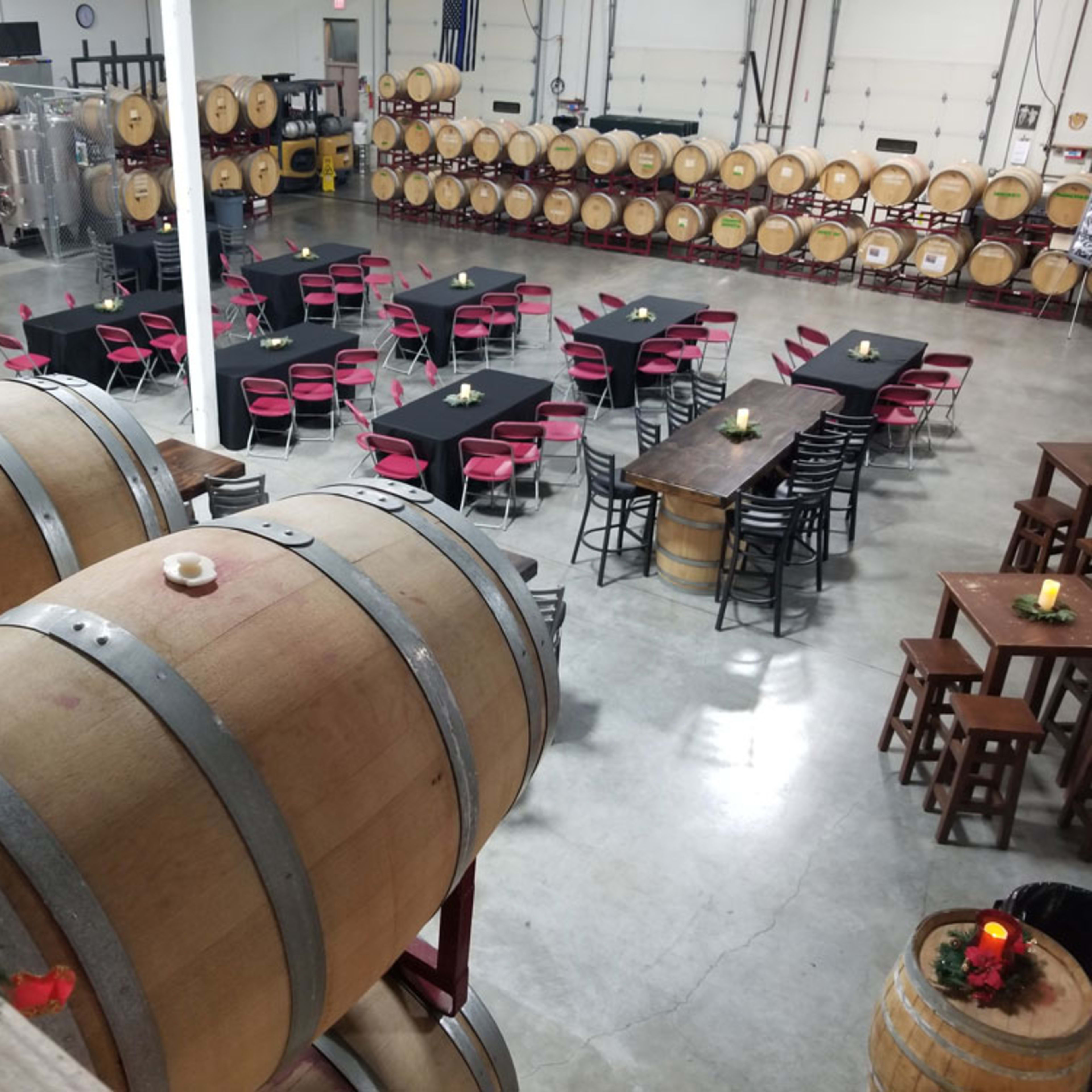 A winery interior with large wooden barrels in the foreground and tables set for dining in the background, surrounded by additional barrels lining the walls.