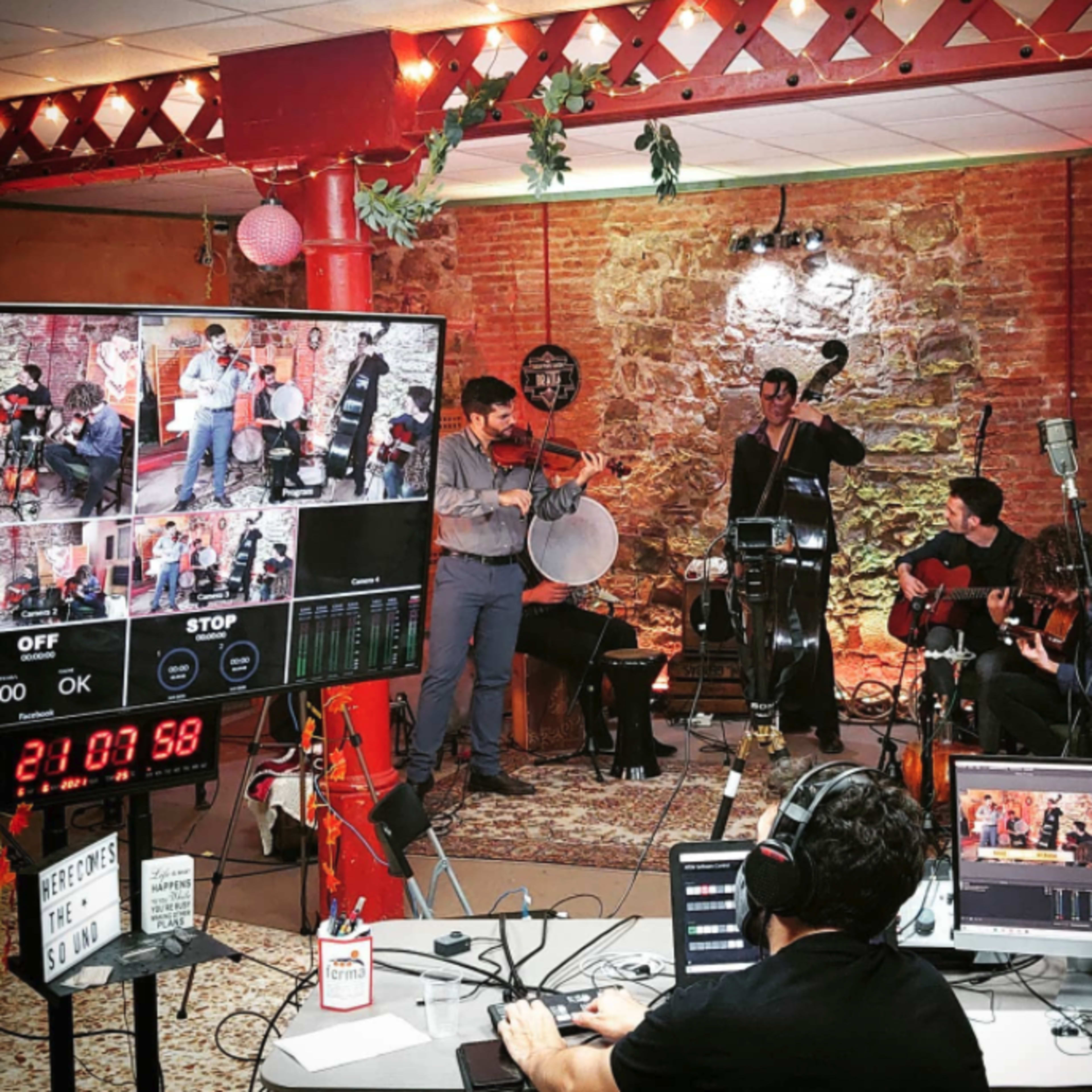 A group of musicians performs in a rustic room equipped with cameras and monitors for a live-streamed event.