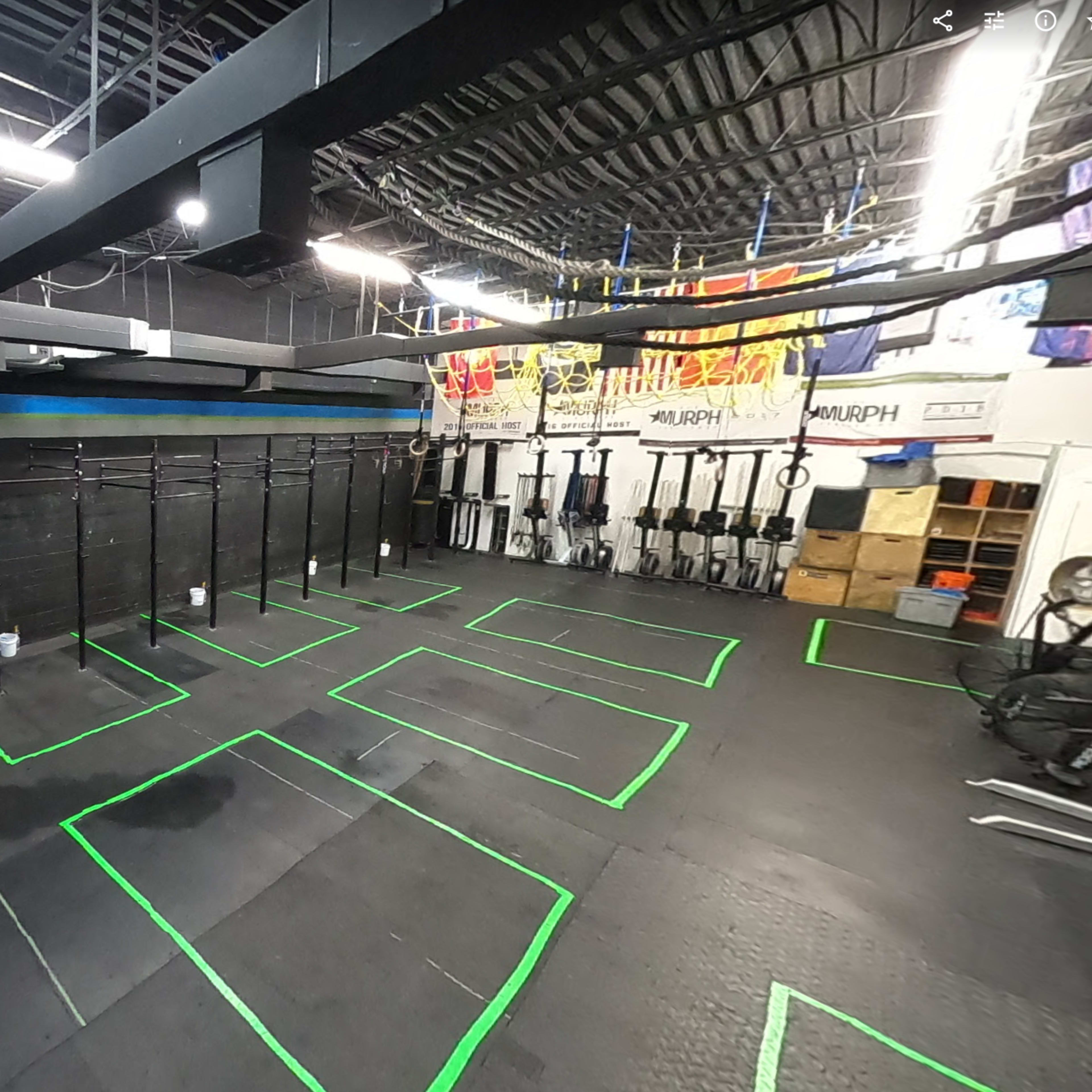 A gym interior with black walls, workout equipment arranged along the back, and green floor markings for exercise stations.