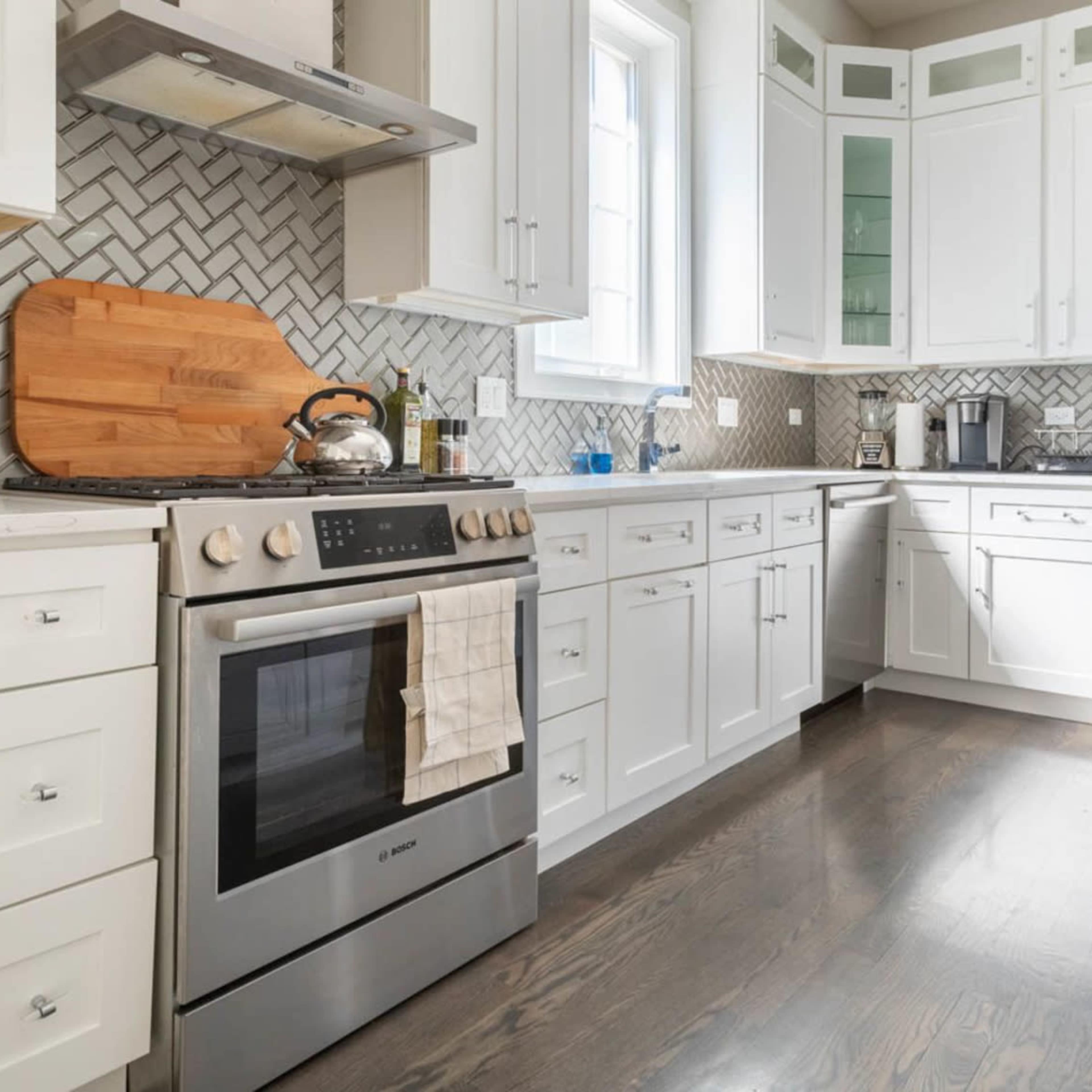 A modern kitchen features white cabinetry, a stainless steel stove with an organized countertop, and a herringbone tiled backsplash.