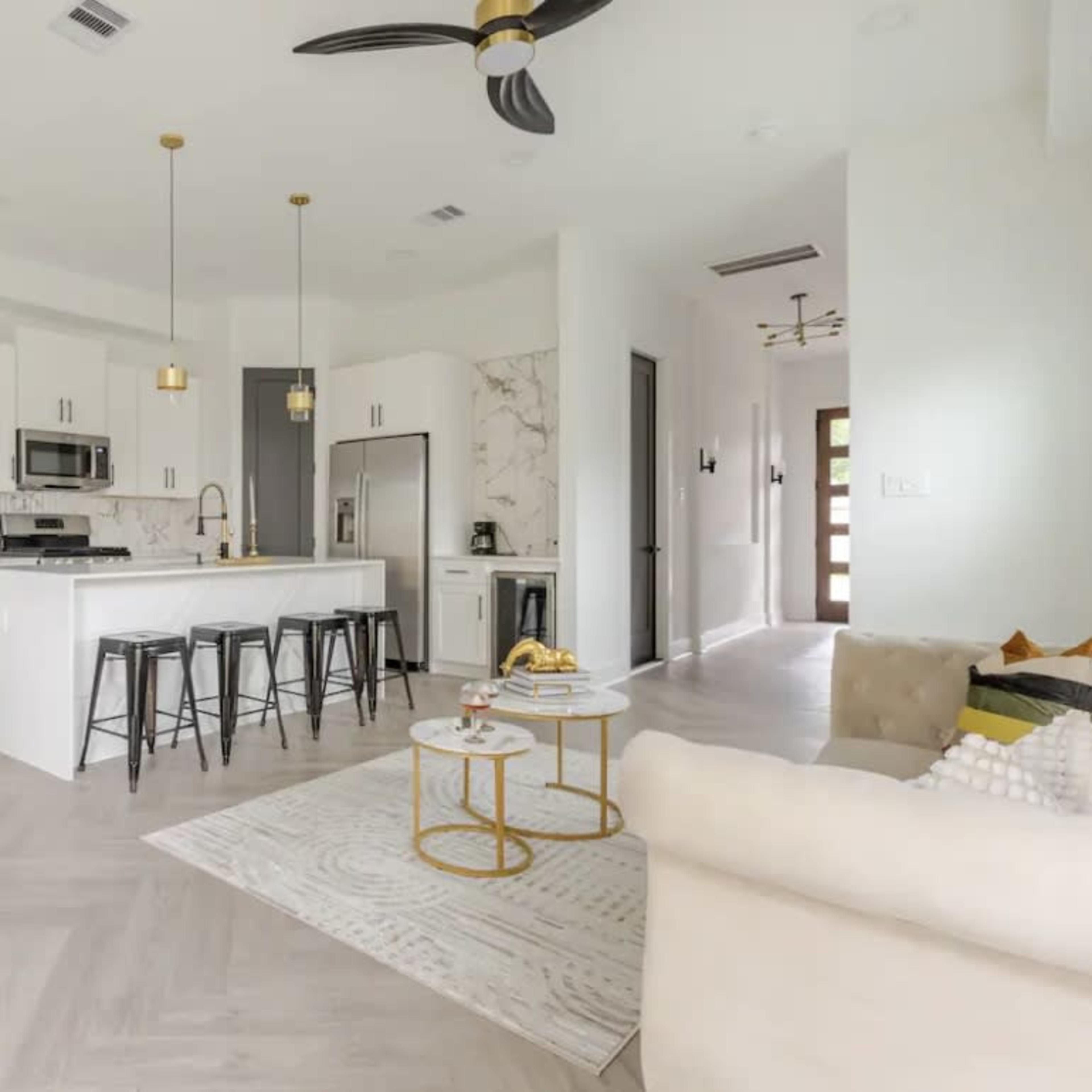 A modern kitchen and living area feature white cabinetry, stainless steel appliances, and a spacious island with bar stools.