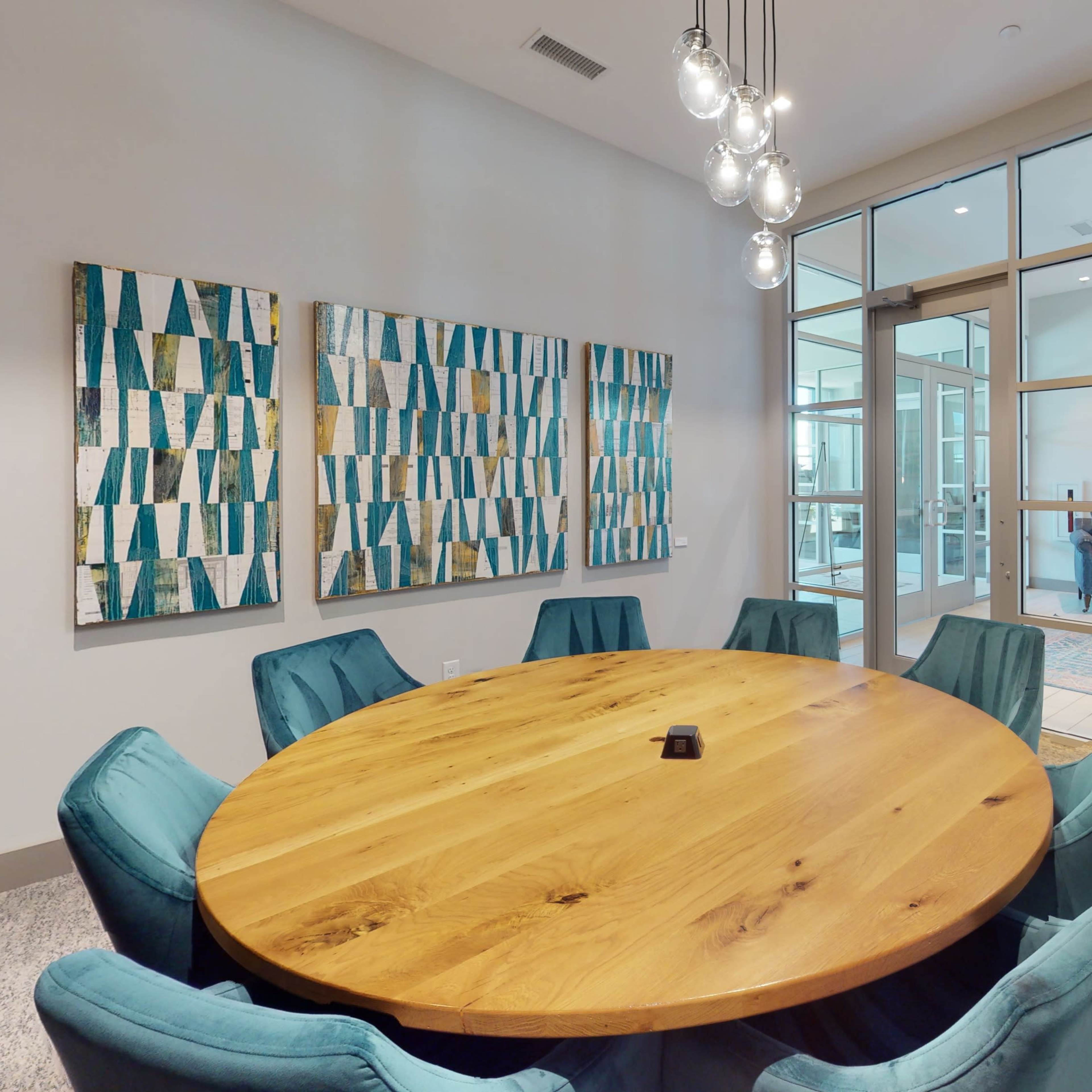 A modern meeting room featuring a large round wooden table surrounded by teal chairs, with abstract artwork on the walls and glass doors leading to a lounge area.