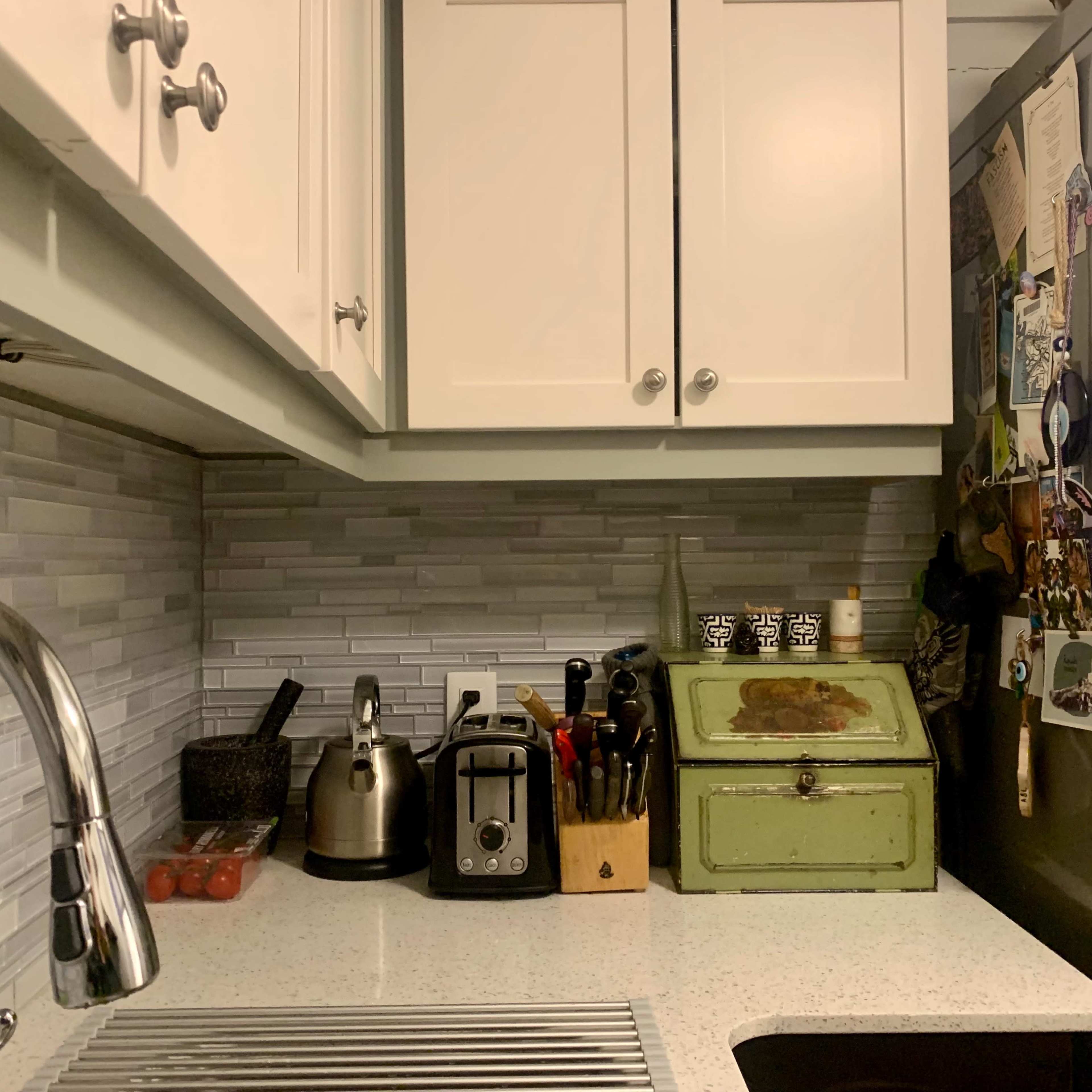 A kitchen countertop with a silver sink, a kettle, a toaster, a set of kitchen utensils, a green storage box, and a fridge adorned with various magnets and notes.