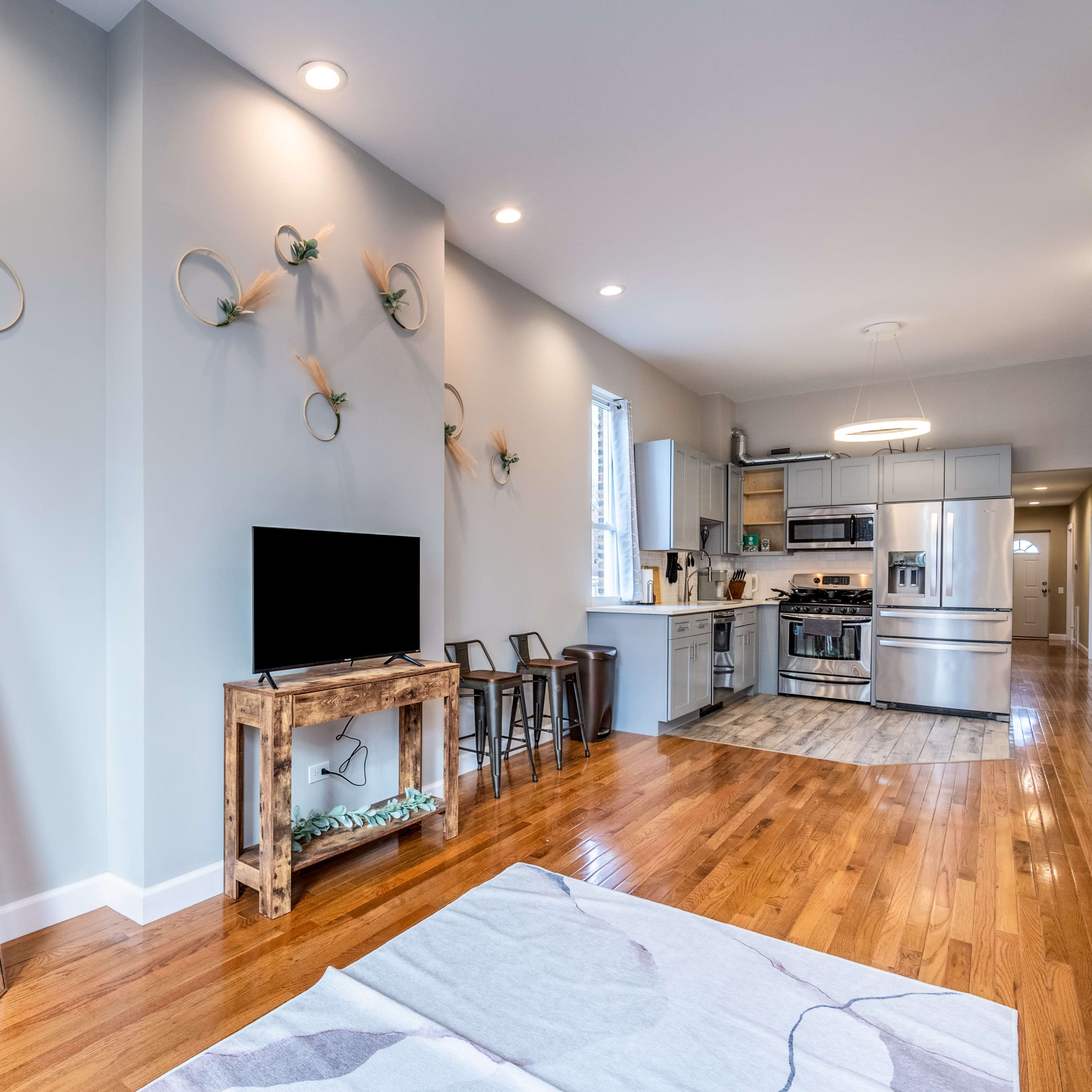 A modern living space featuring a kitchen area, wooden flooring, a television on a wooden stand, and decorative wall accents.