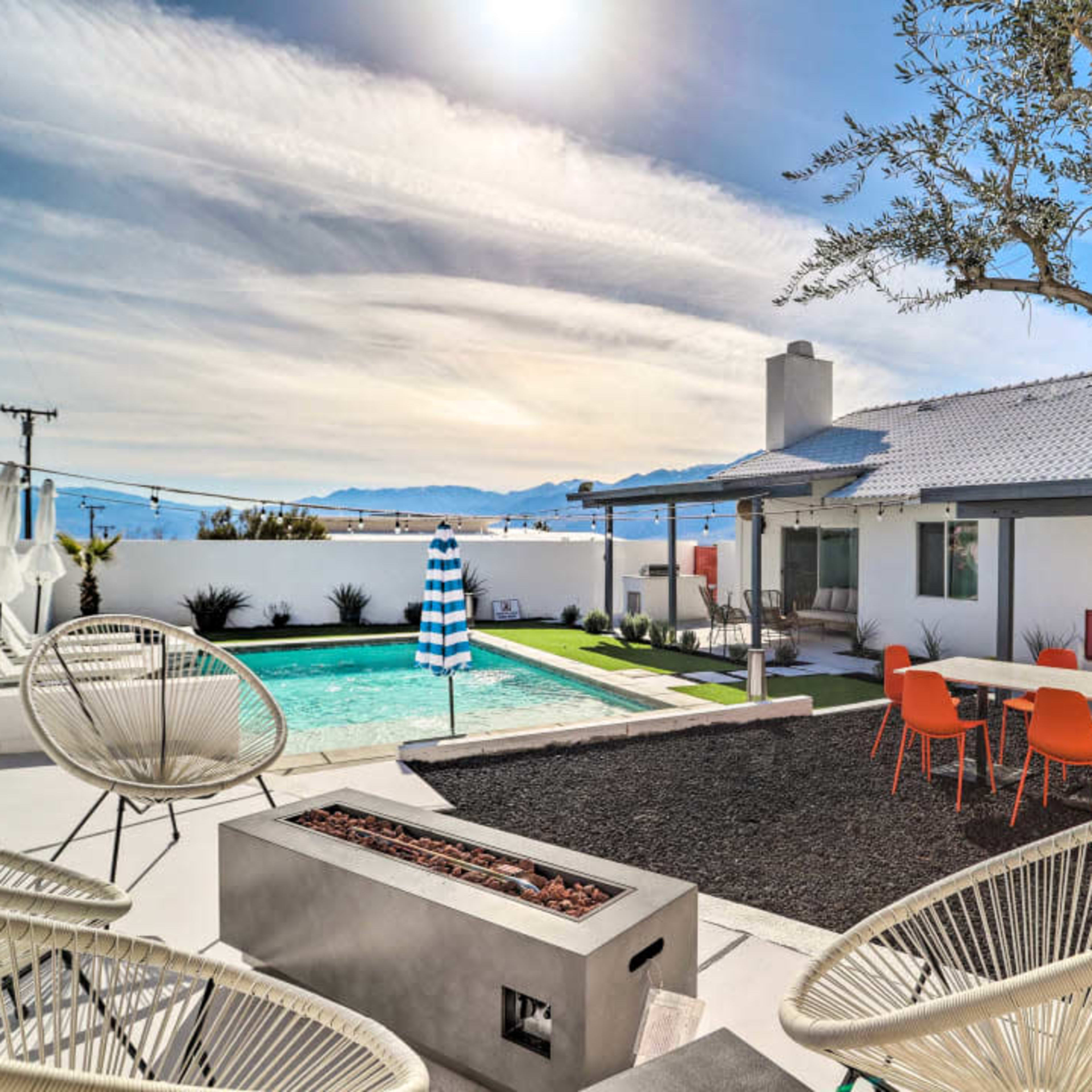 A modern outdoor space with a swimming pool, patio furniture, and a landscaped area surrounded by mountains under a cloudy sky.