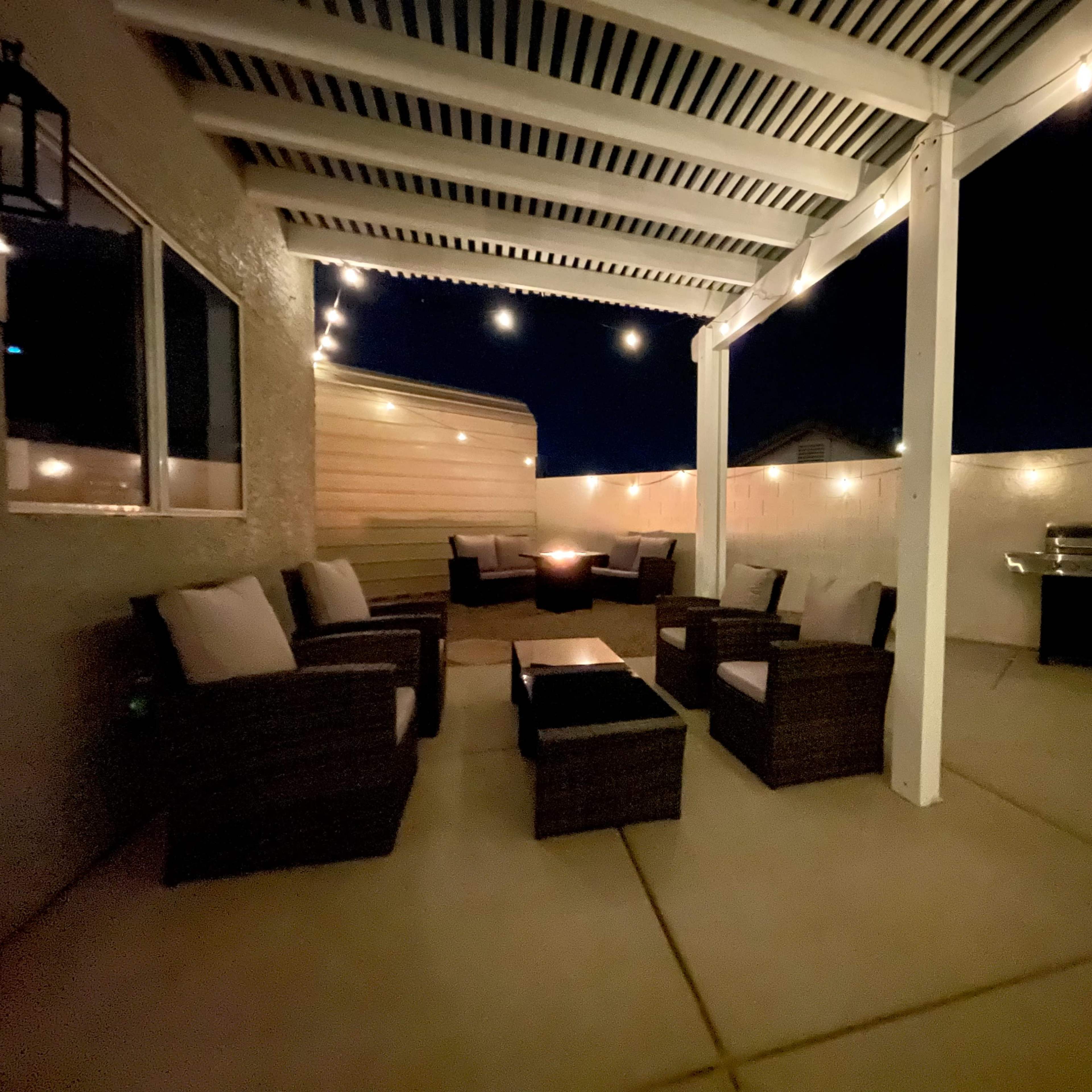 A patio area with wicker seating, a fire pit, and a grill, illuminated by string lights at night.