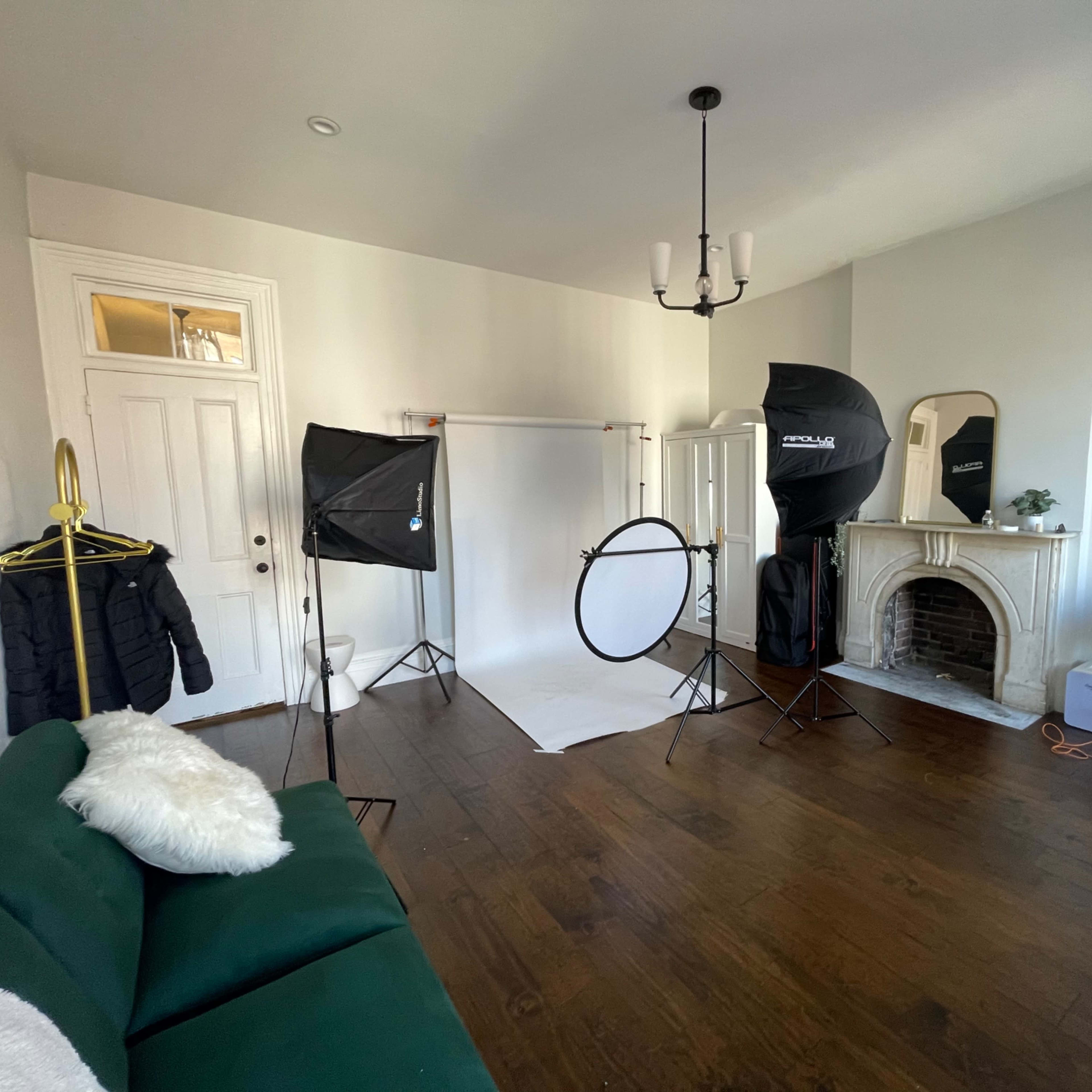 A well-lit room set up for photography, featuring a green couch, backdrop stands, light modifiers, and a mirror near an ornate fireplace.