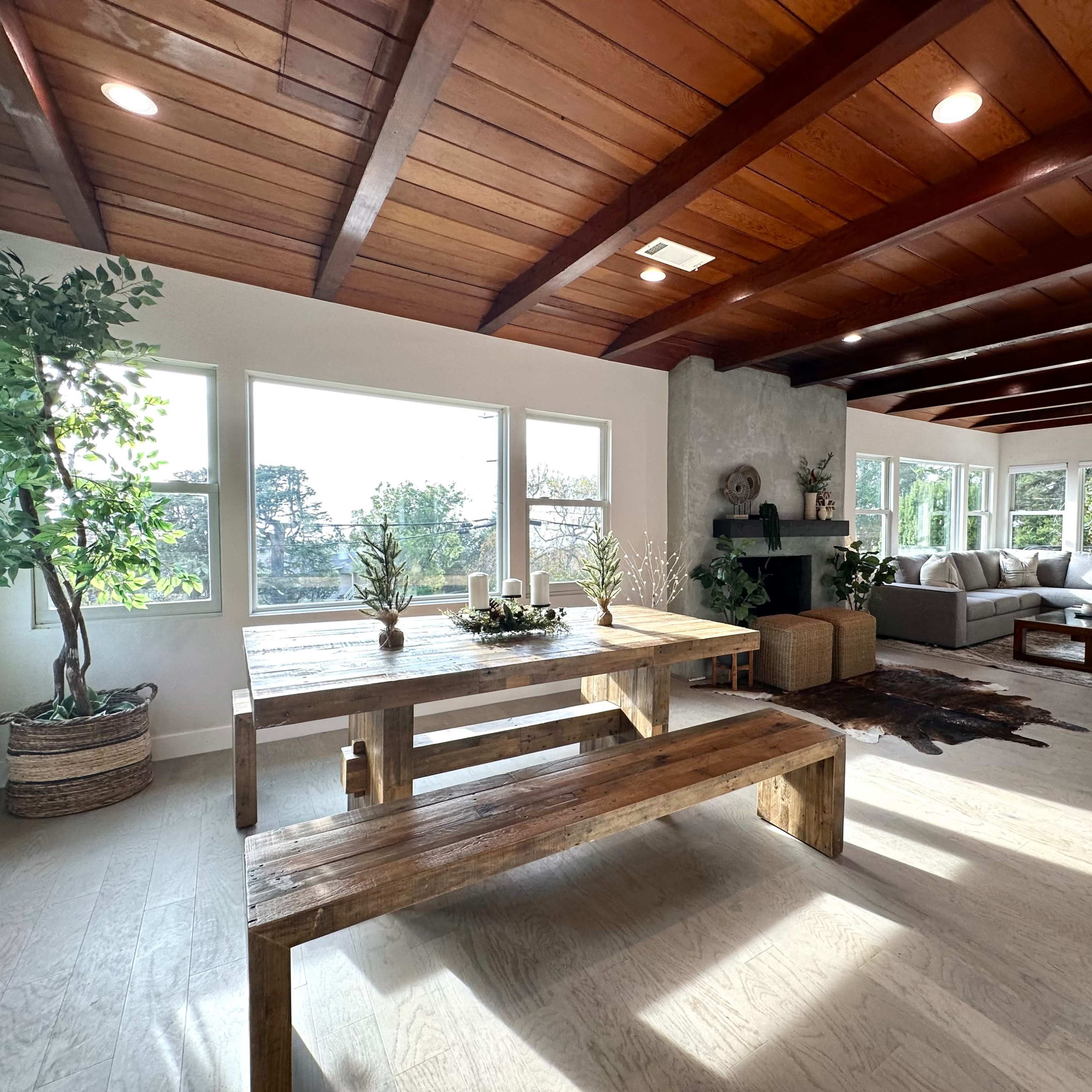 A bright, open living area featuring a wooden table with benches, a gray sofa, and large windows overlooking greenery.