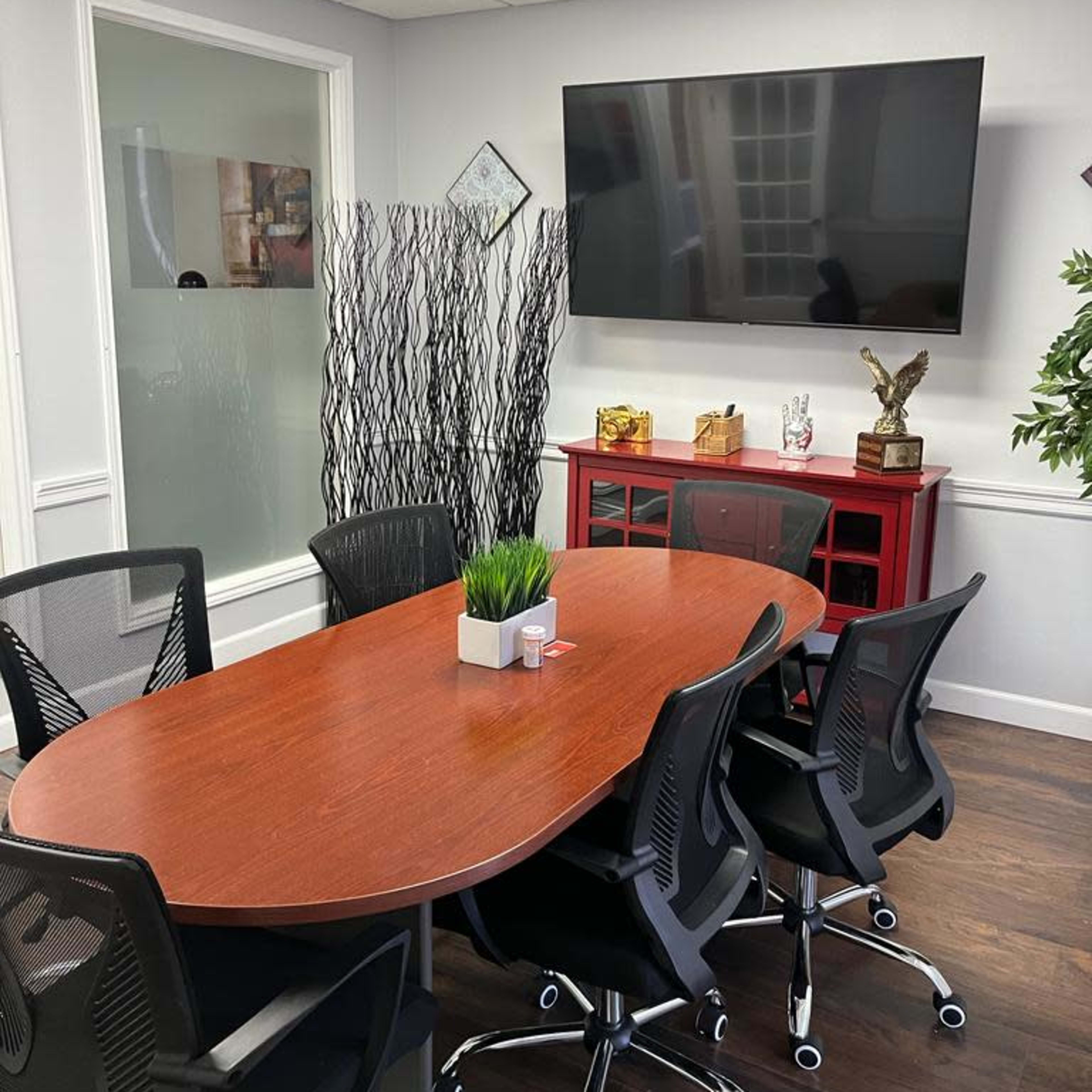 A modern conference room featuring a wooden oval table surrounded by black mesh chairs, a large television mounted on the wall, and decorative elements including a plant and a divider.