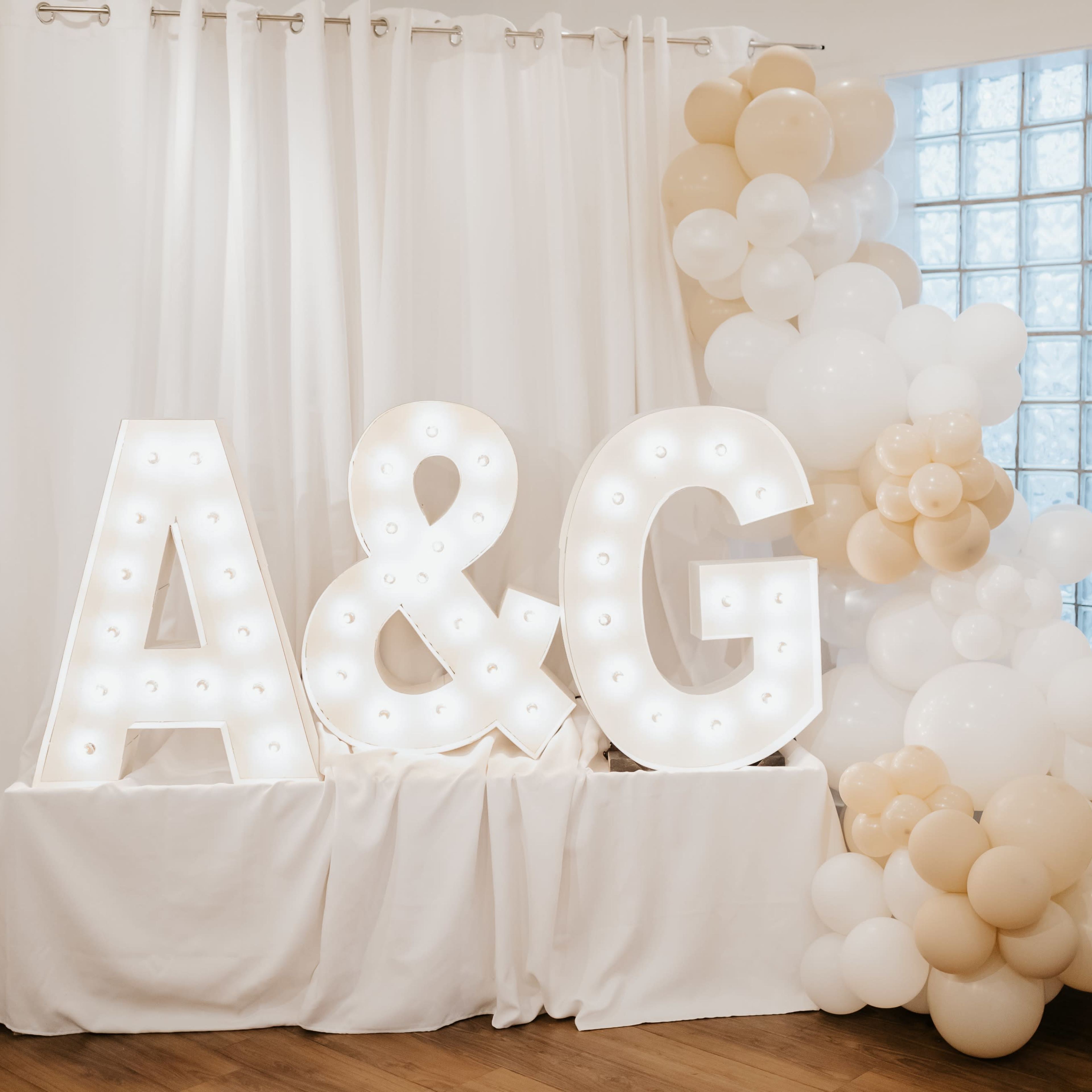 Illuminated letters "A" and "G" against a white curtain, accompanied by a decorative arrangement of balloons in neutral tones.