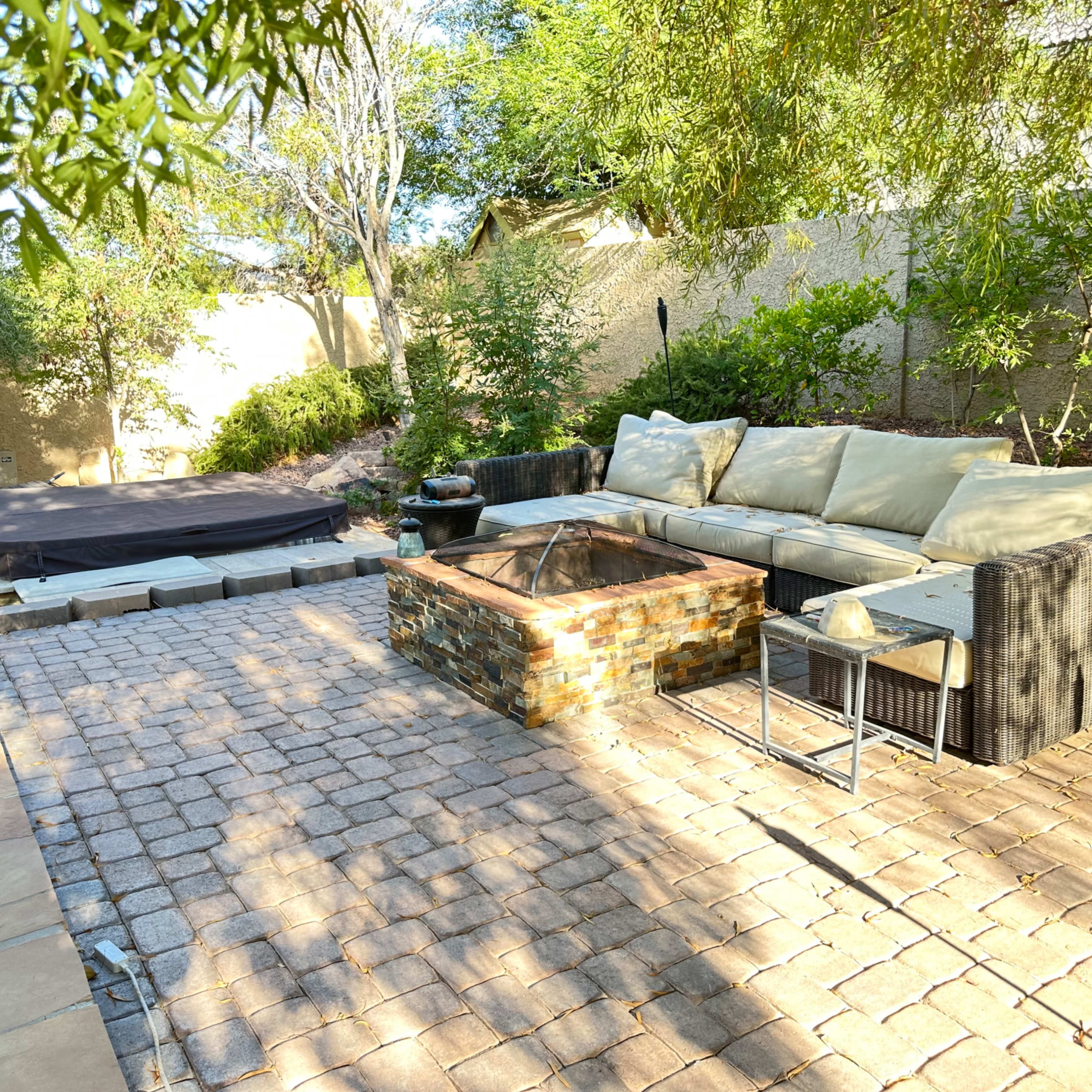 The outdoor patio features a stone firepit surrounded by a sectional sofa and a covered hot tub in a landscaped garden.