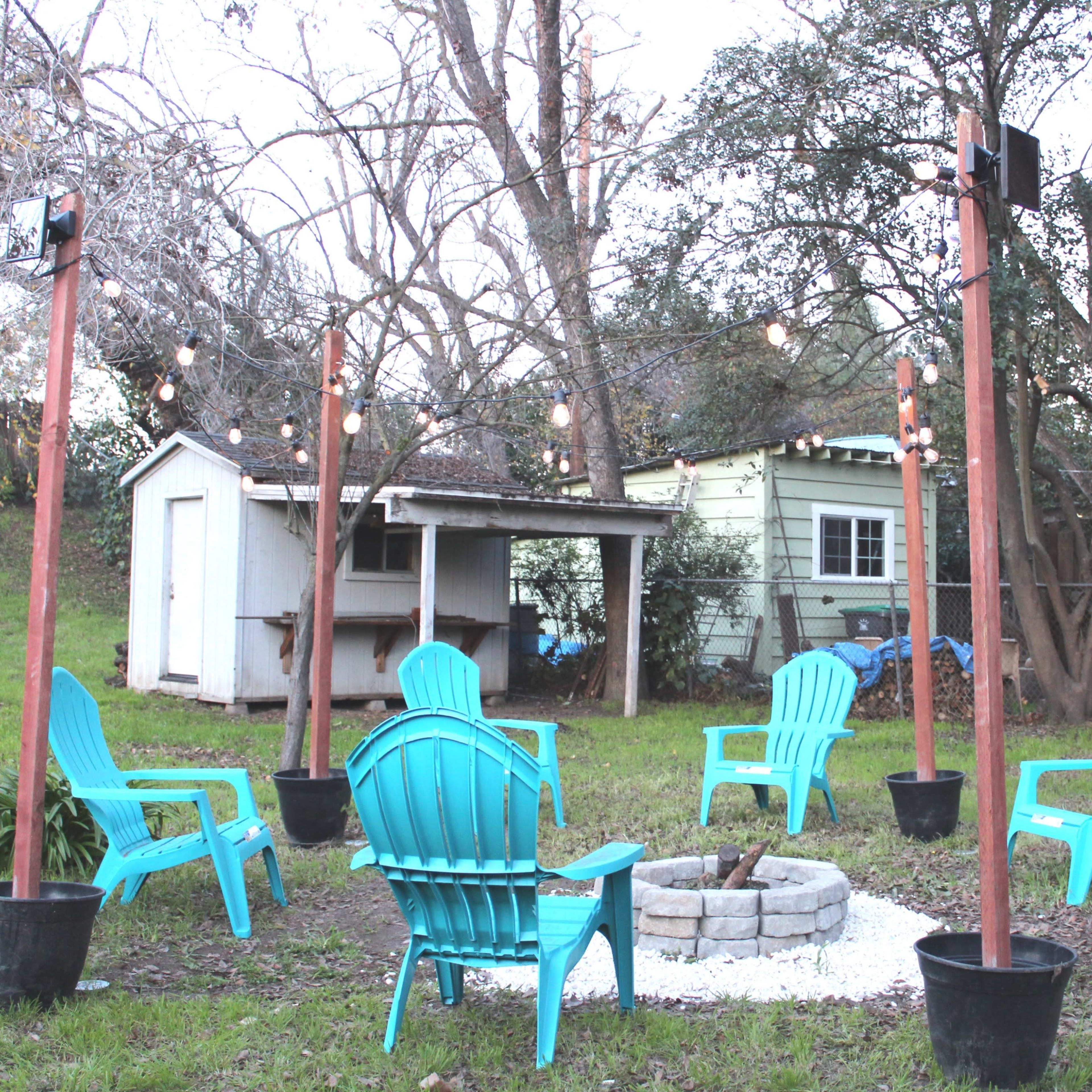A circular gathering space with blue adirondack chairs surrounds a stone fire pit, with string lights overhead and a shed in the background.
