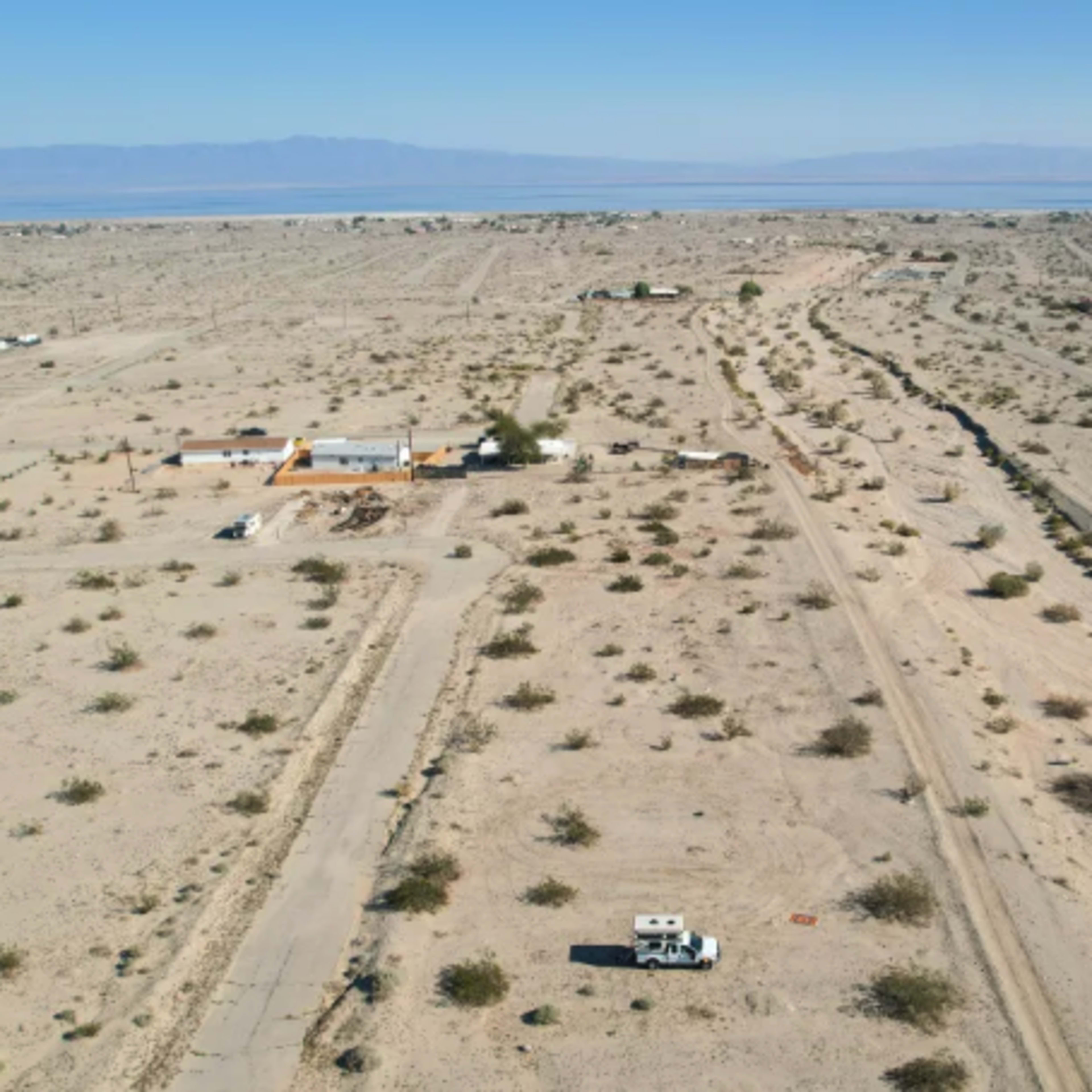 A barren desert landscape with sparse vegetation, scattered buildings, and an RV parked on a dirt road.