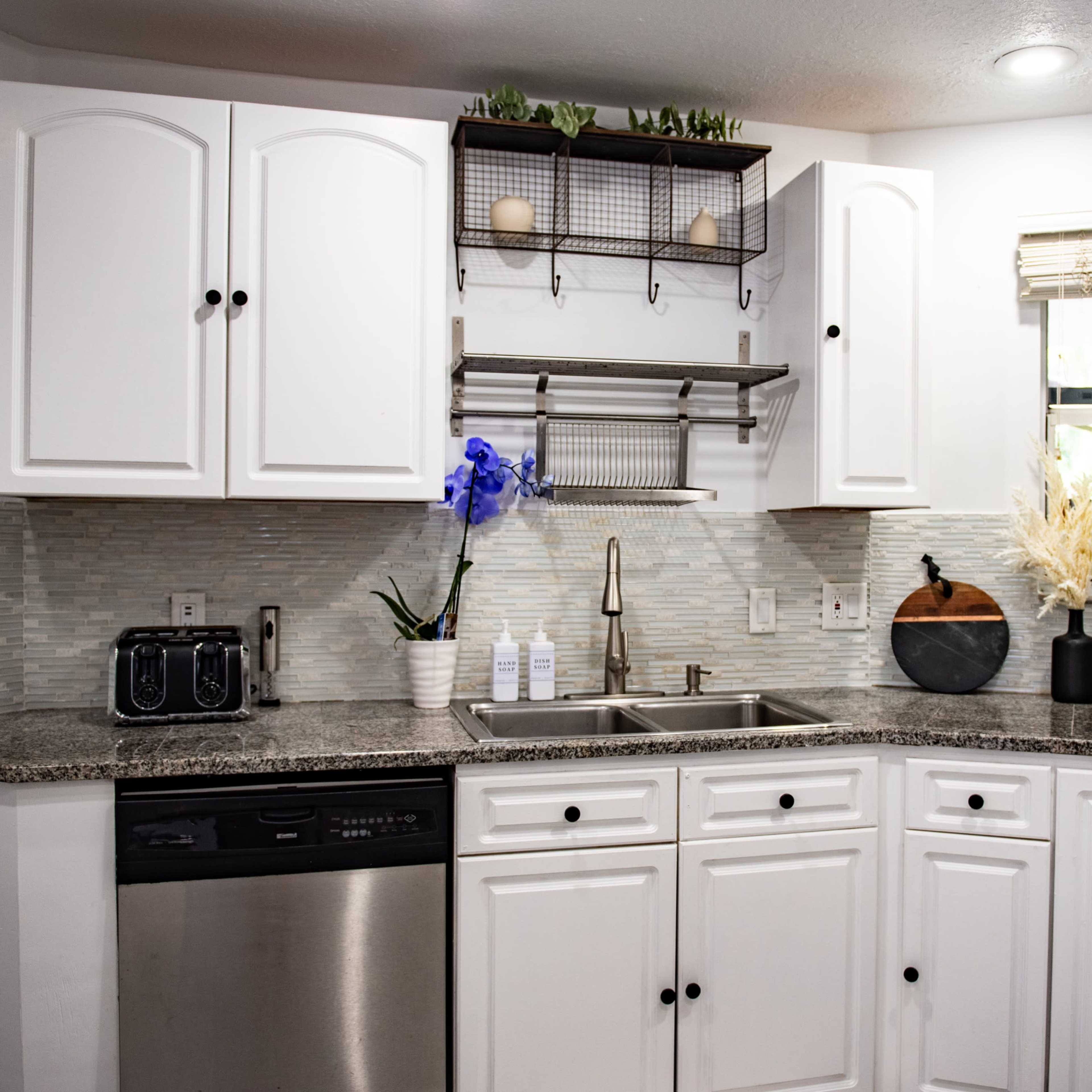 A modern kitchen with white cabinets, a granite countertop, and a stainless steel sink, complete with a small window above and decorative items on the shelves.