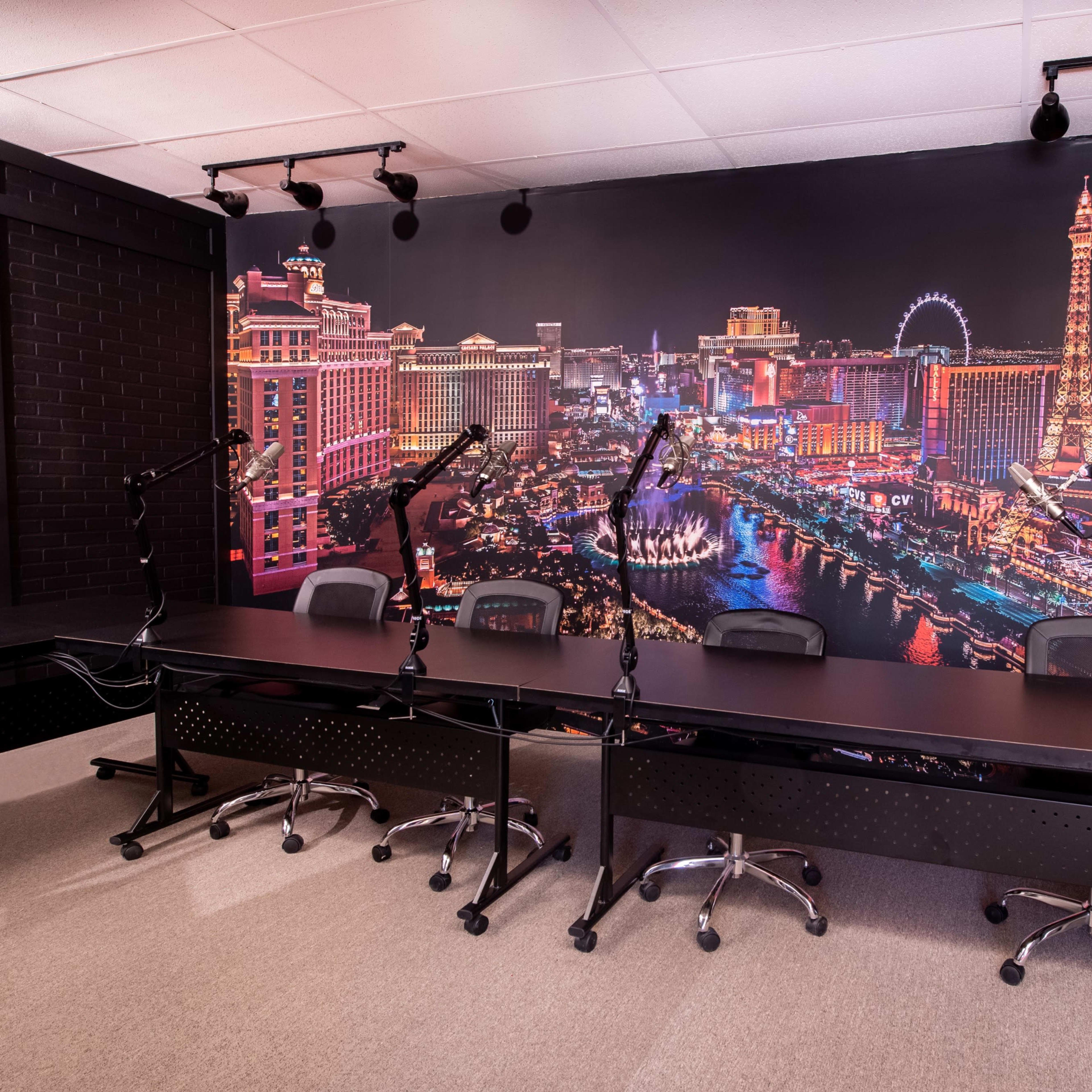 A podcast studio featuring a long table with several chairs, multiple microphone stands, and a large backdrop of the Las Vegas skyline illuminated at night.