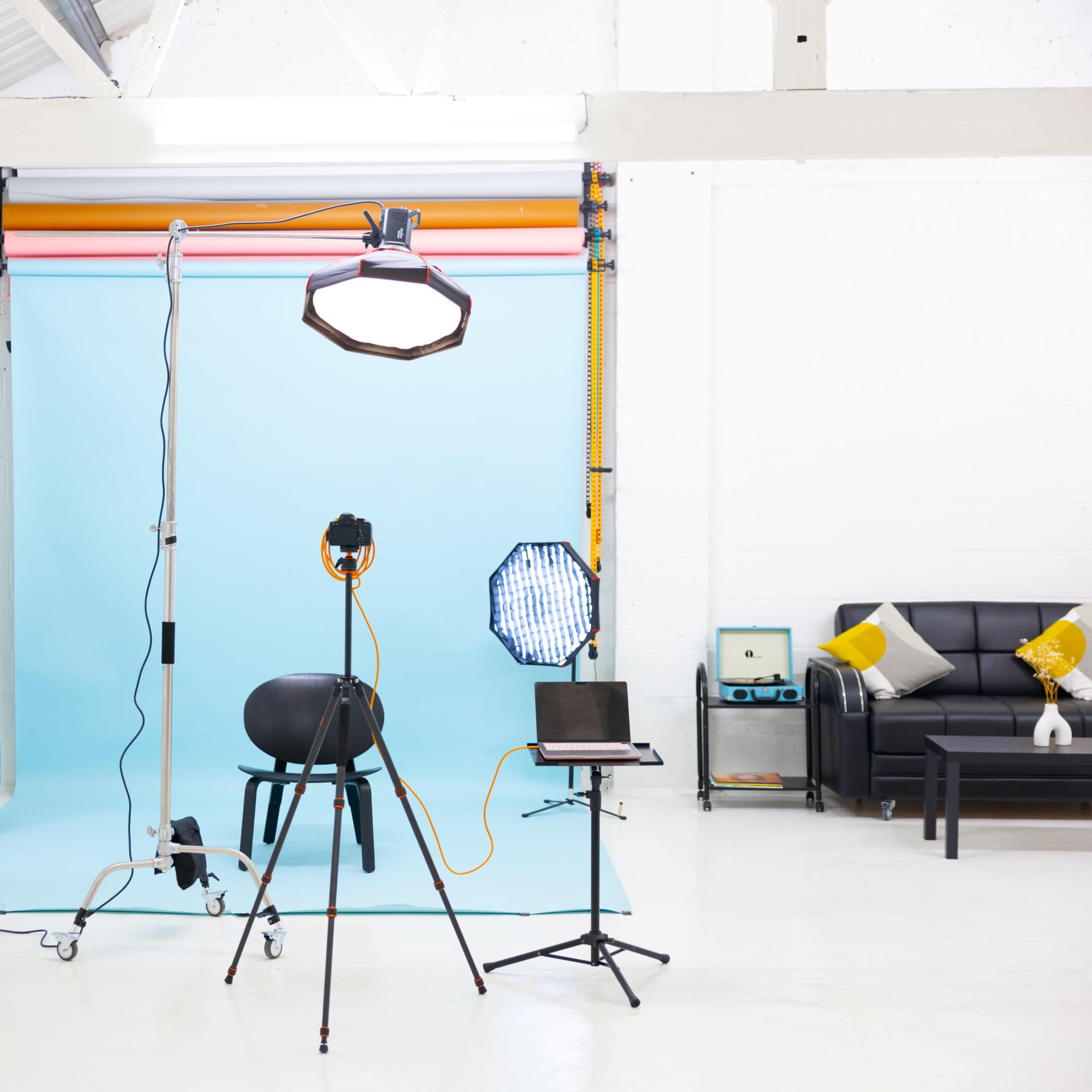 A photography studio features lighting equipment, a blue backdrop, a black chair, and a small table with chairs.