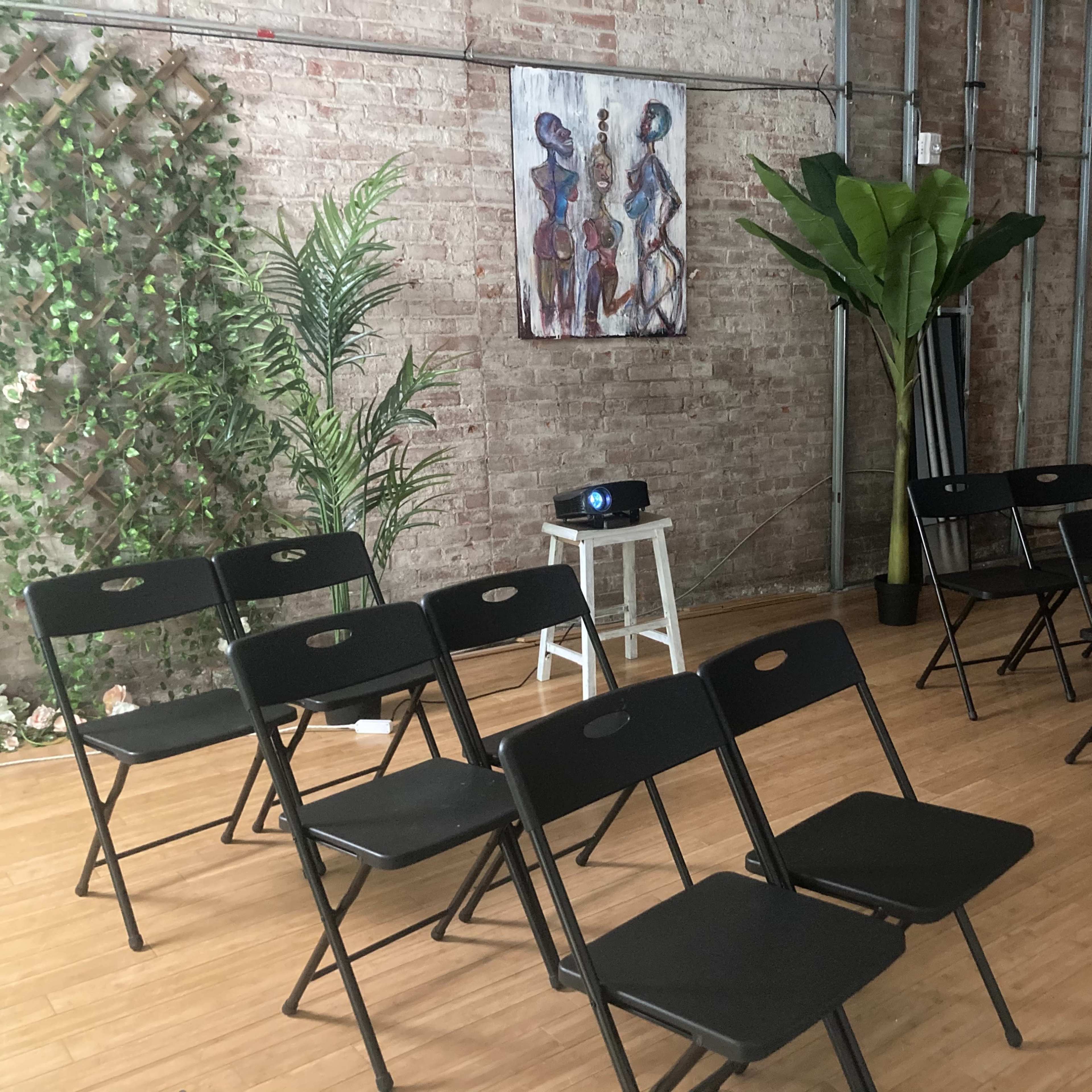 A room with a brick wall, featuring several black folding chairs arranged in rows, a small projector on a white stool, and a painting on the wall alongside a potted plant.