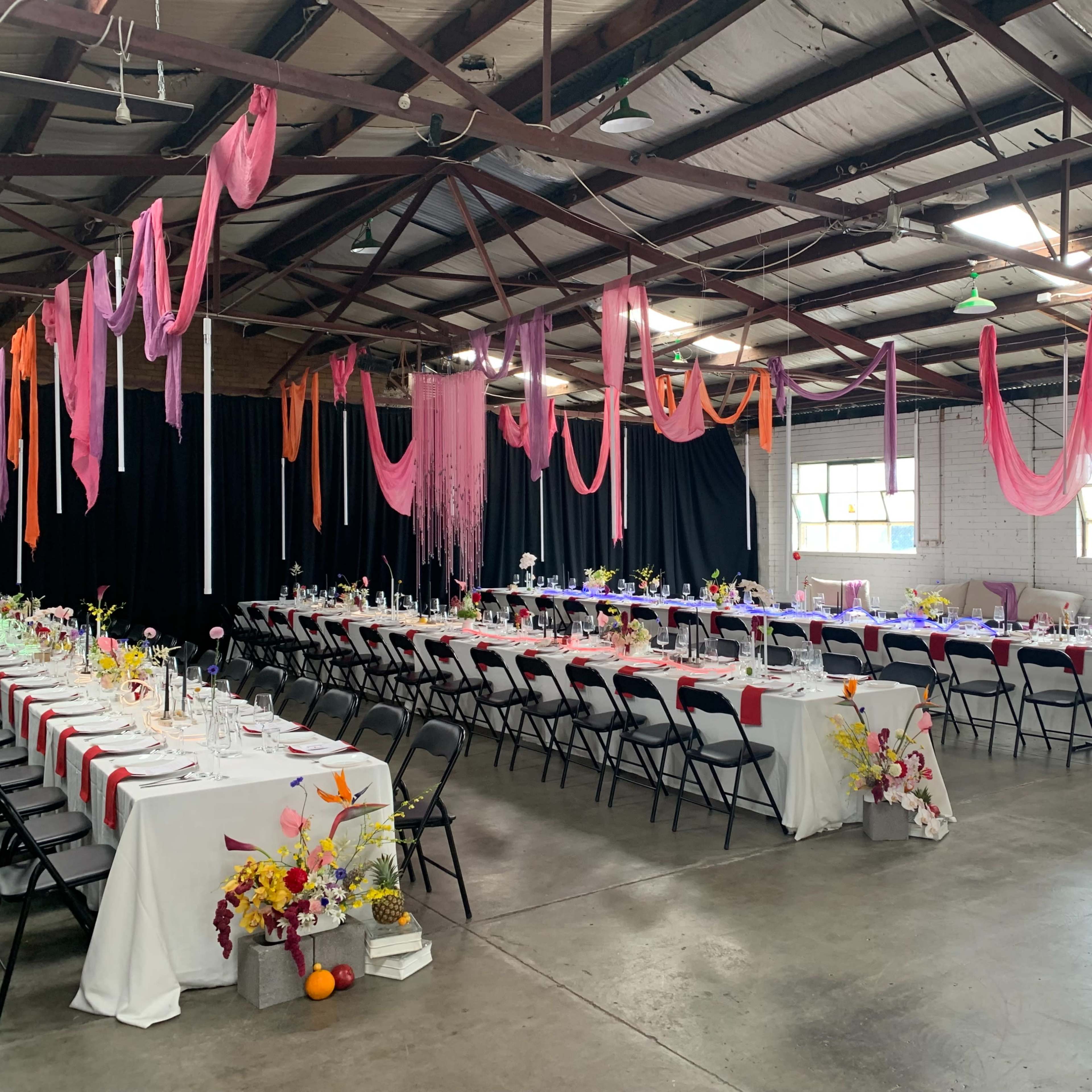 A spacious indoor venue is decorated with colorful drapery and floral arrangements, featuring long banquet tables and surrounding chairs.