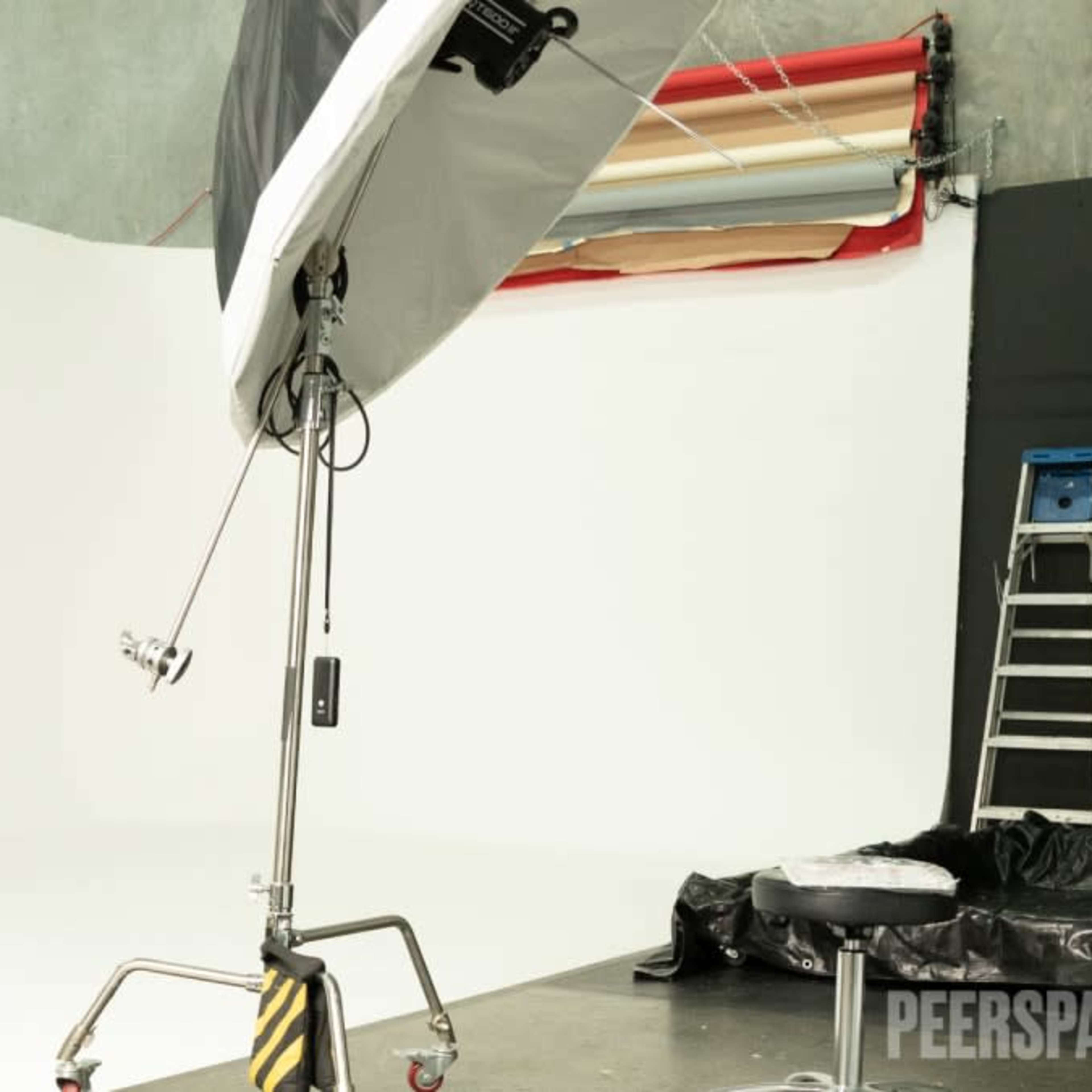 A photography studio features a large softbox light setup next to a blank white backdrop and a ladder.