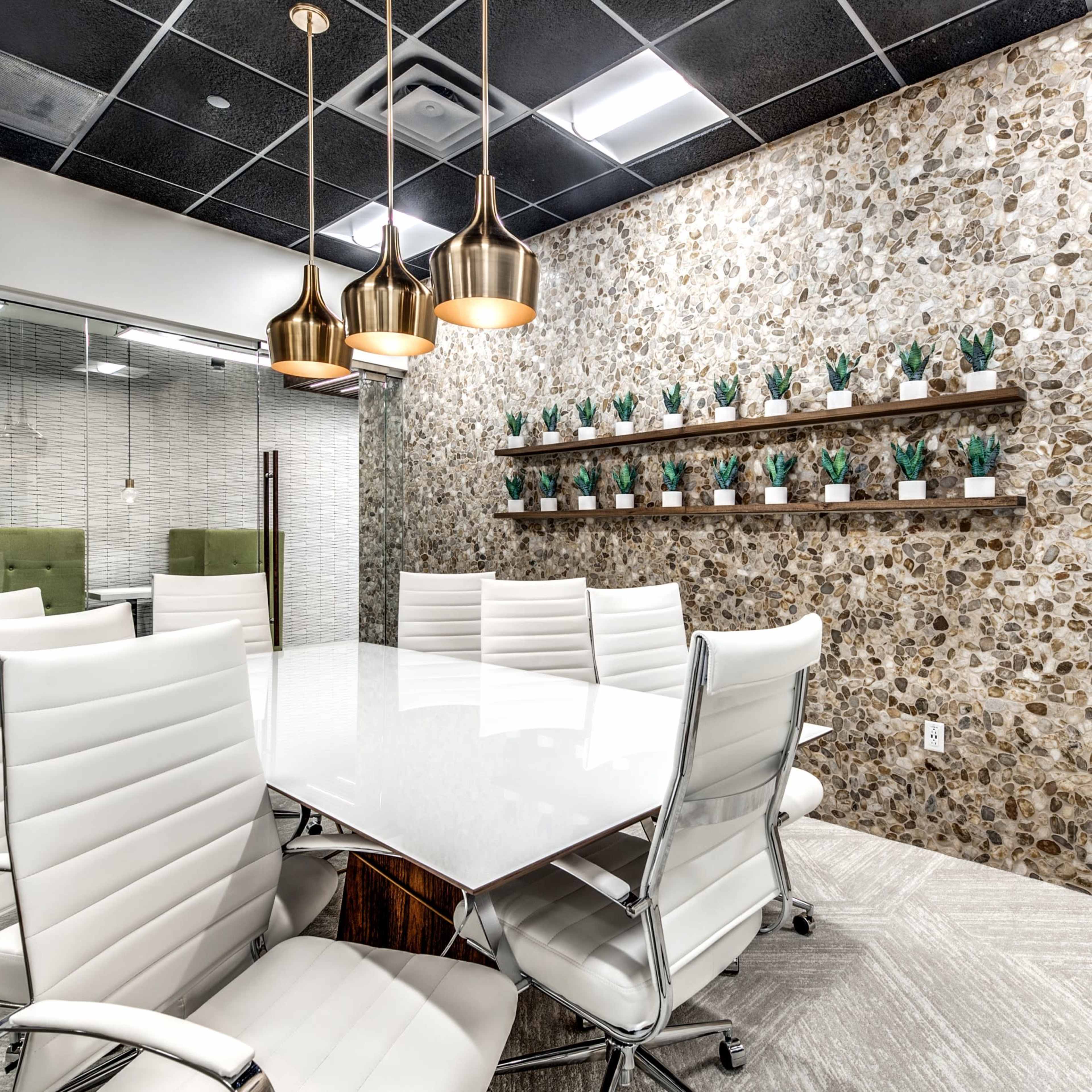 A modern conference room features a white marble table surrounded by sleek white chairs, with a stone accent wall decorated with small potted plants and pendant lighting overhead.