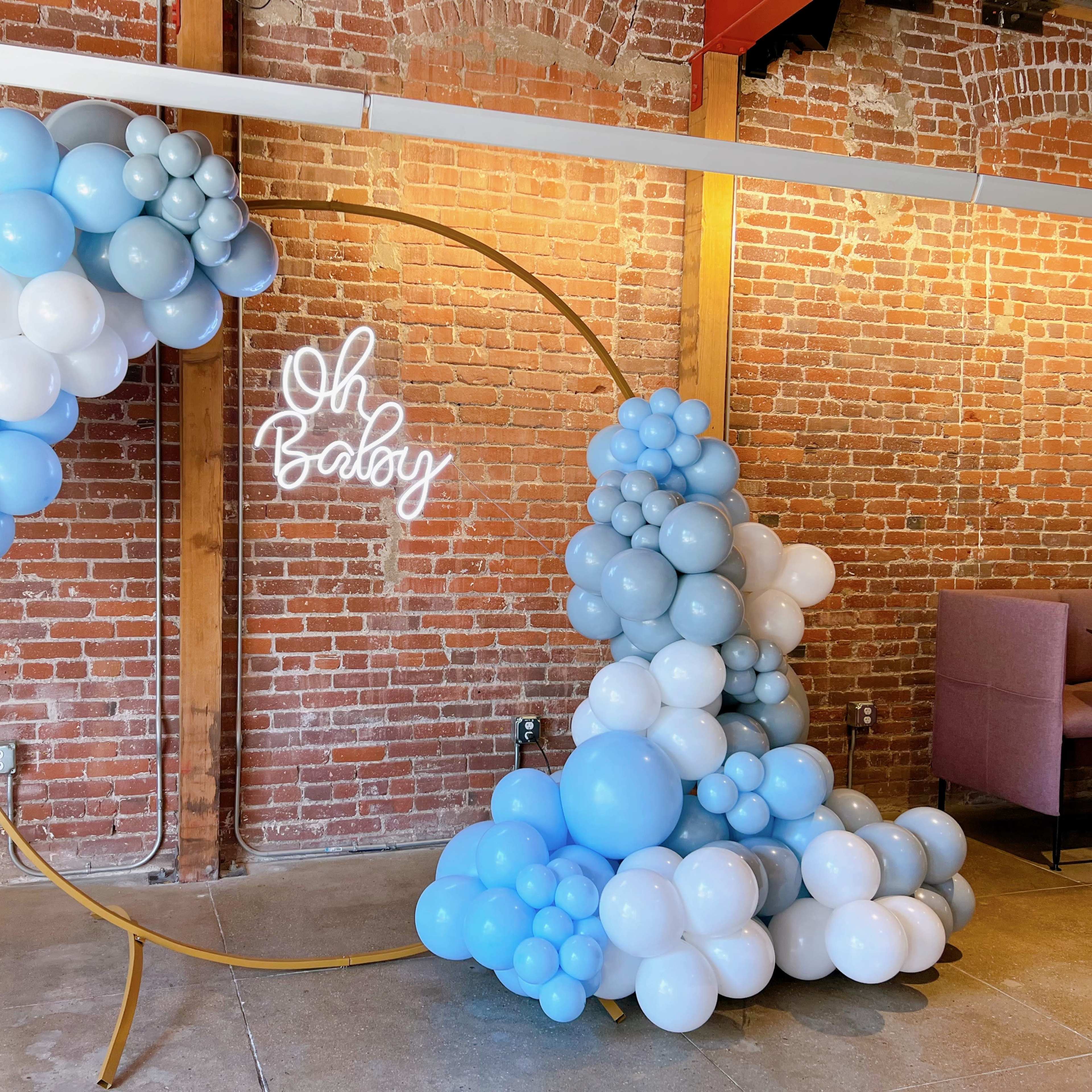 A circular balloon arch composed of blue and gray balloons, positioned against a backdrop of exposed brick, with a neon sign that reads "Oh Baby."