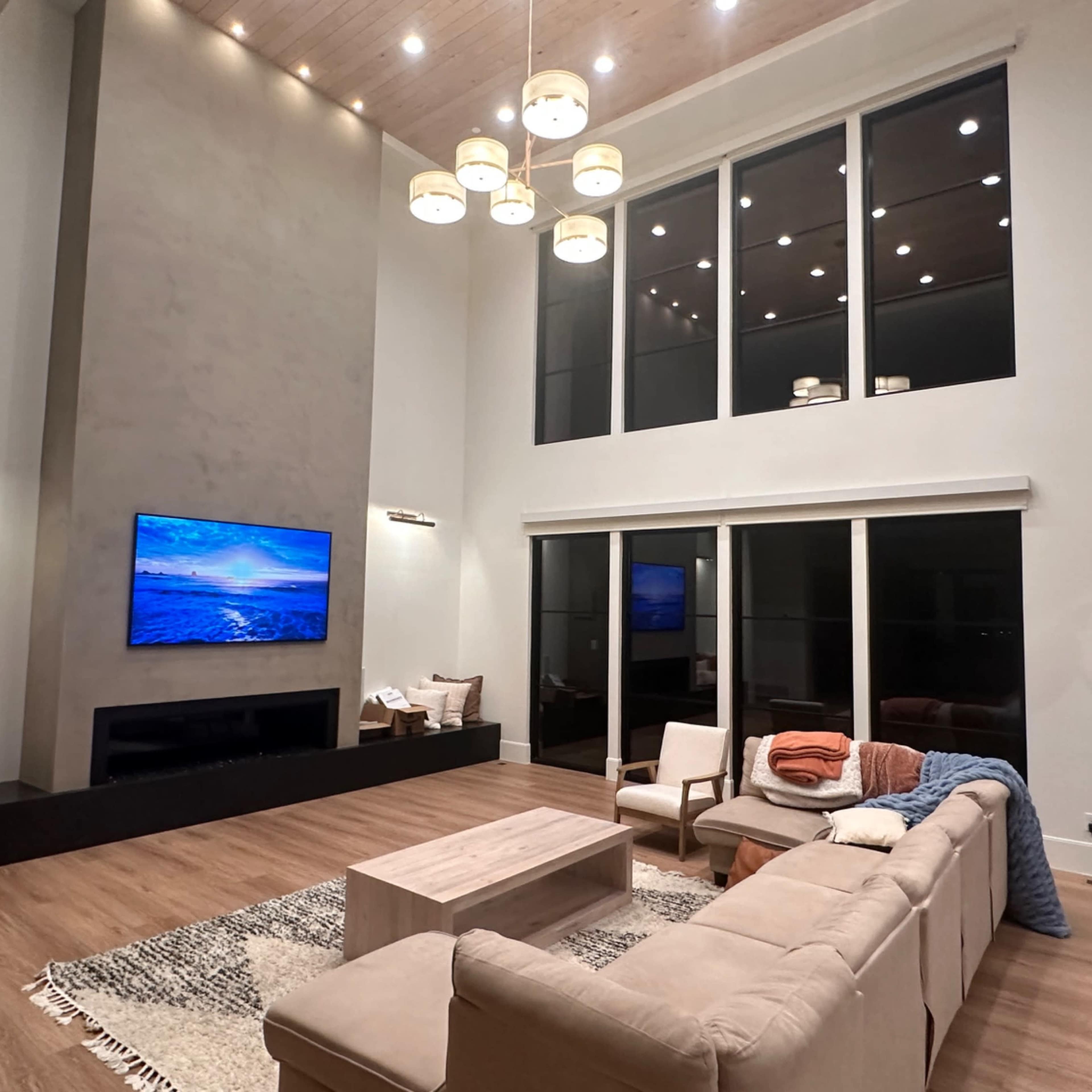 A modern living room features a large sectional sofa, a coffee table, a wall-mounted television displaying a scenic view, large windows, and a high ceiling adorned with multiple light fixtures.