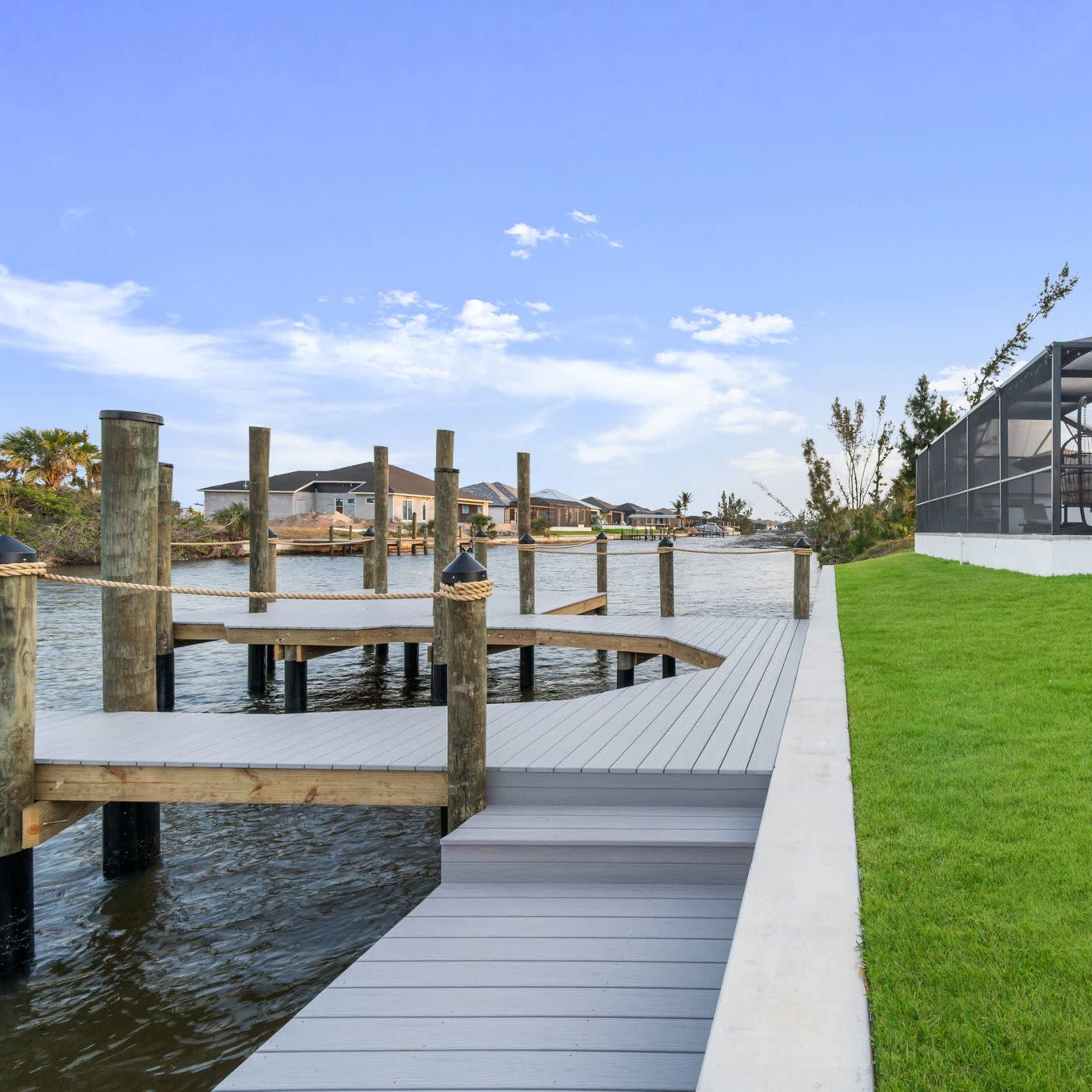 A wooden dock extending into a body of water, with a green lawn and a screened patio house in the background.