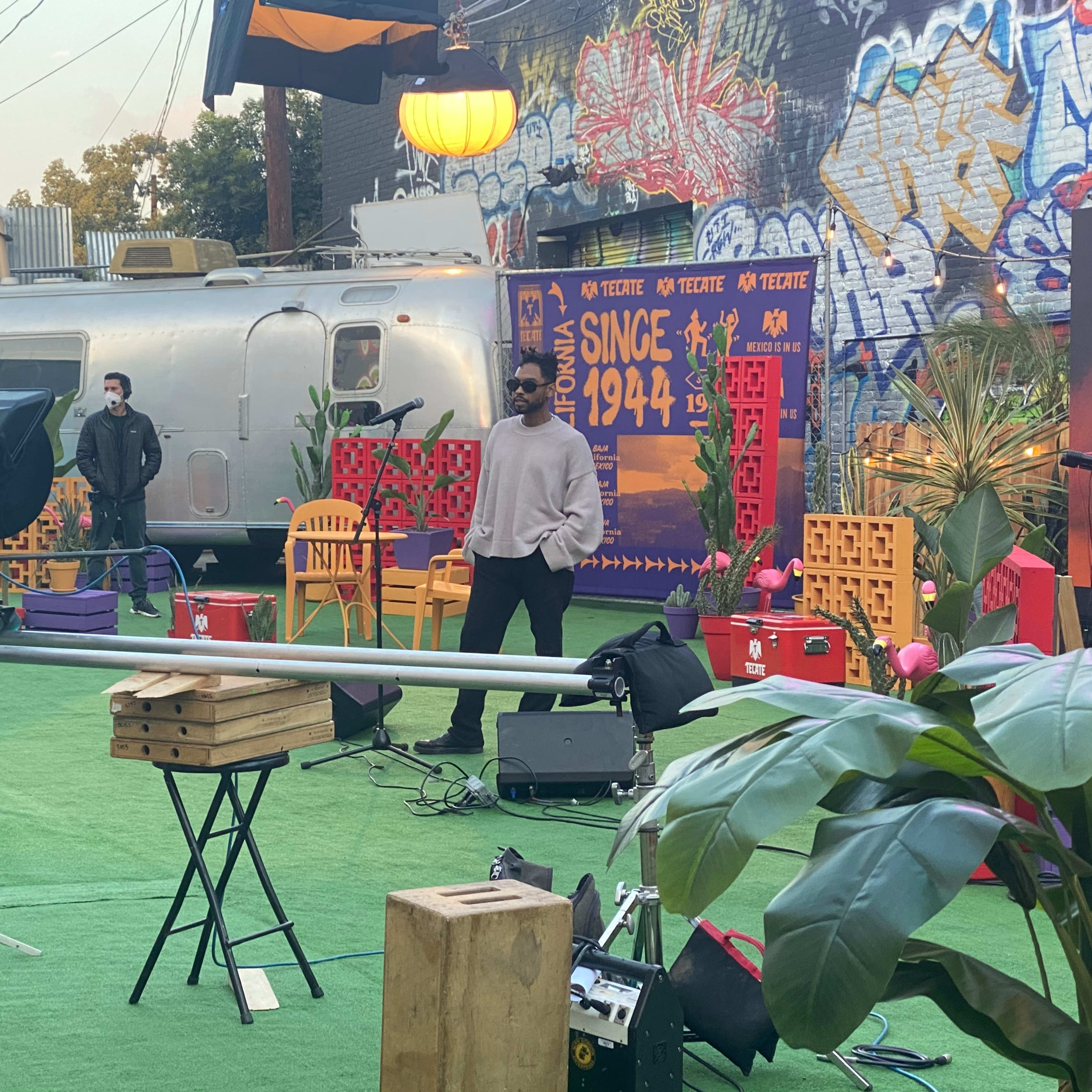 A film crew sets up equipment in an outdoor area featuring colorful decor and a vintage trailer.