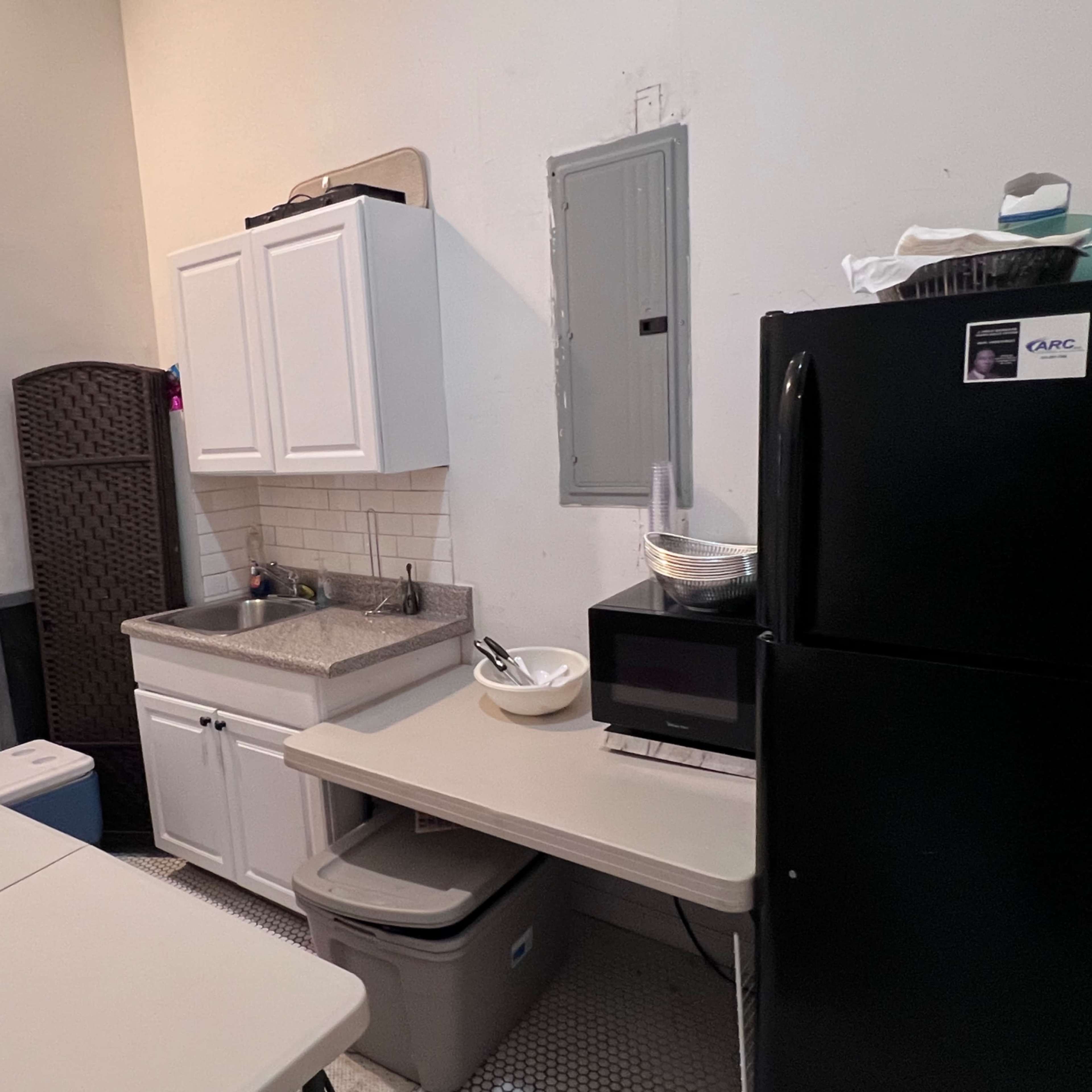 A small kitchen area with a sink, cabinets, a microwave, and a black refrigerator, alongside a table and storage bins.