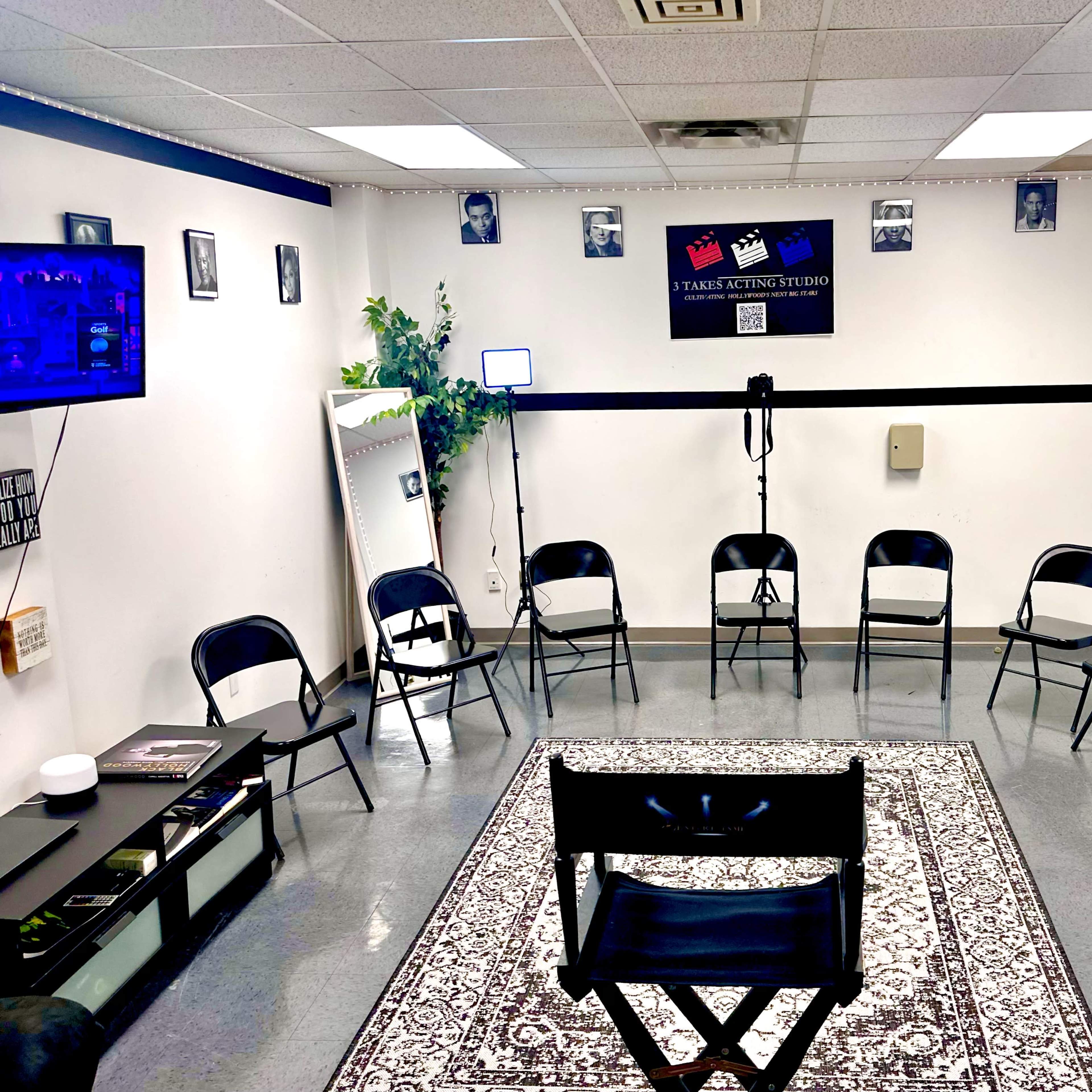 A room set up for a meeting, featuring a circle of black chairs around a central black director's chair, with framed photographs on the walls and a television on one side.