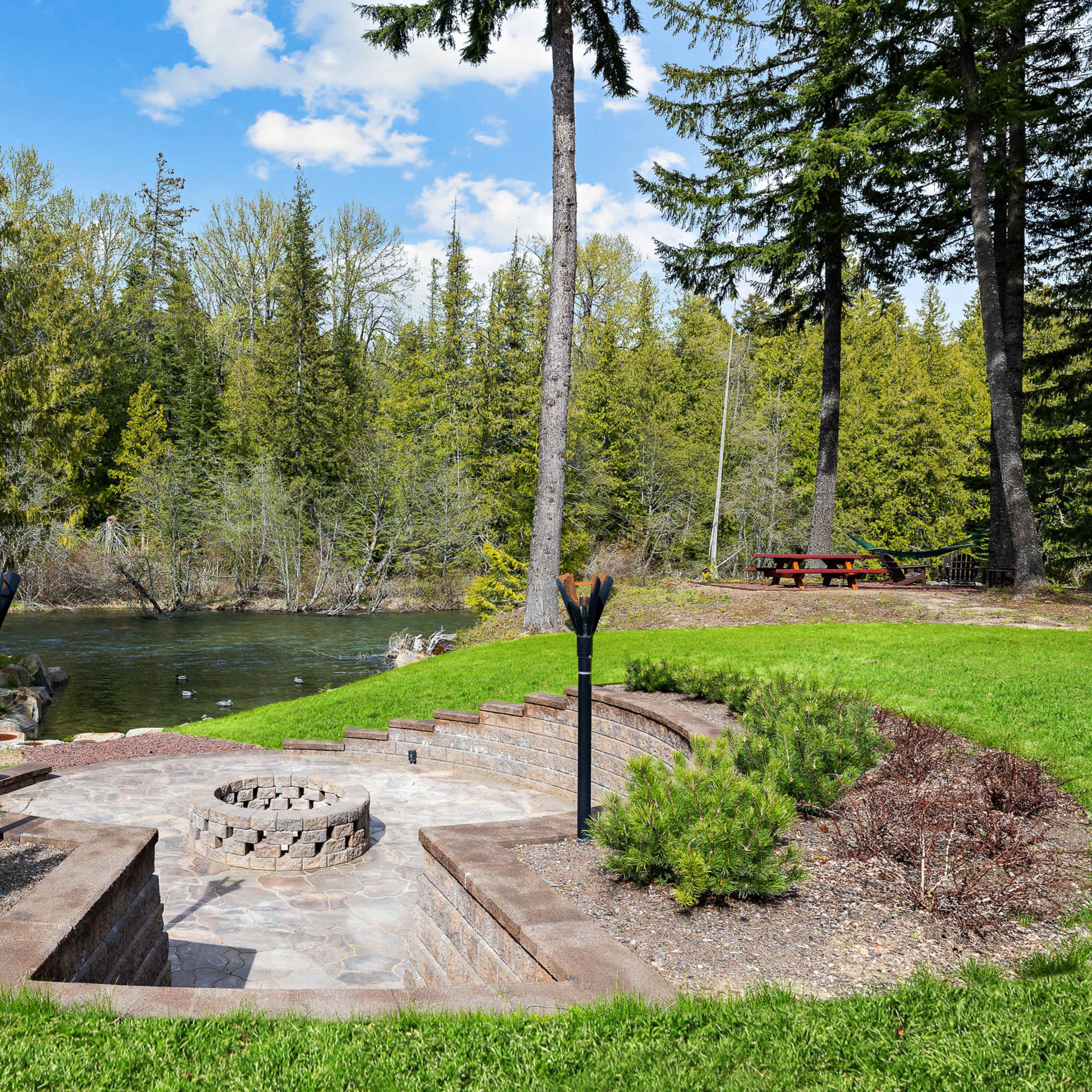A stone patio with a circular fire pit overlooks a calm pond surrounded by trees and a picnic area.