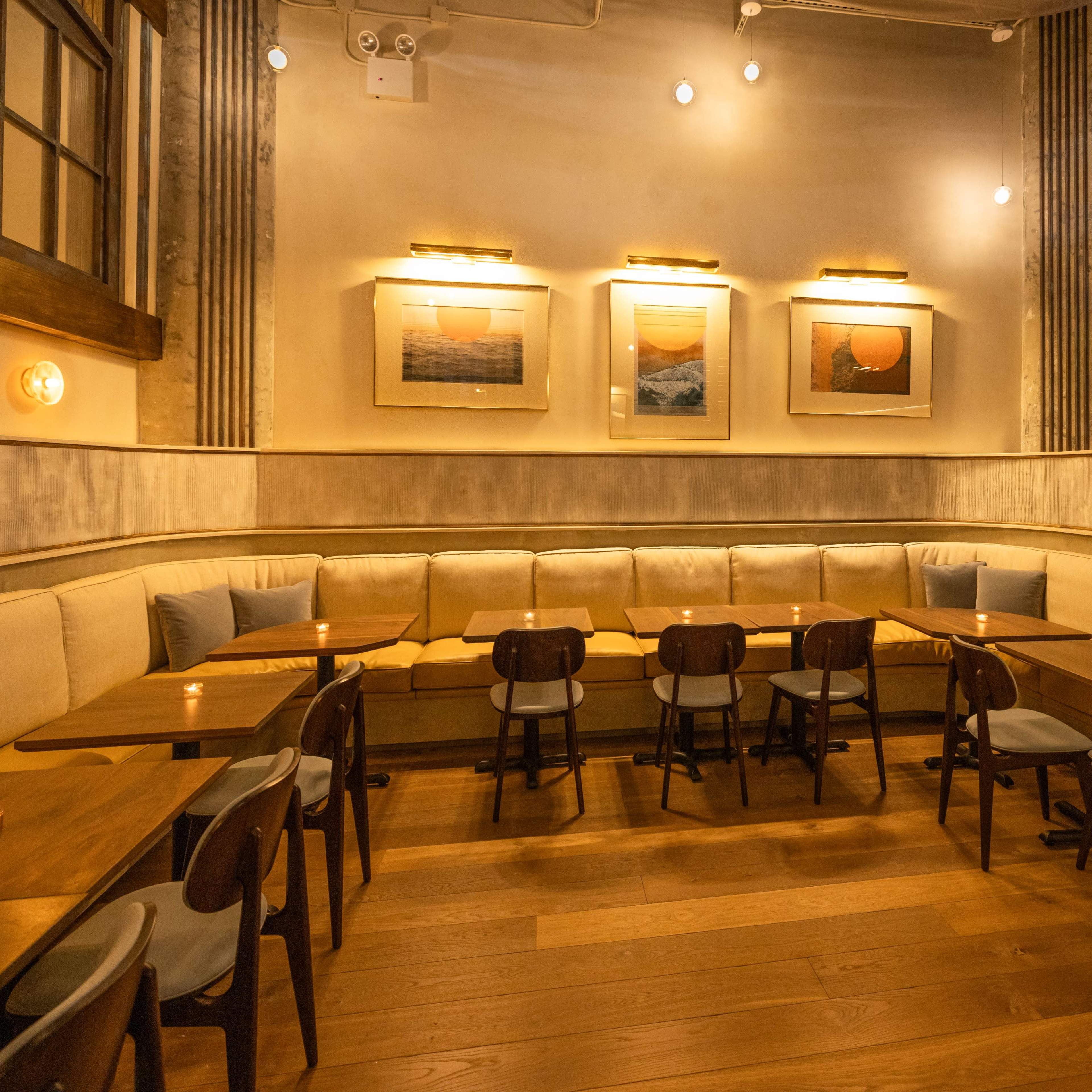 A cozy restaurant interior with wooden tables and chairs arranged around a padded bench along the wall, featuring framed artwork and soft lighting.