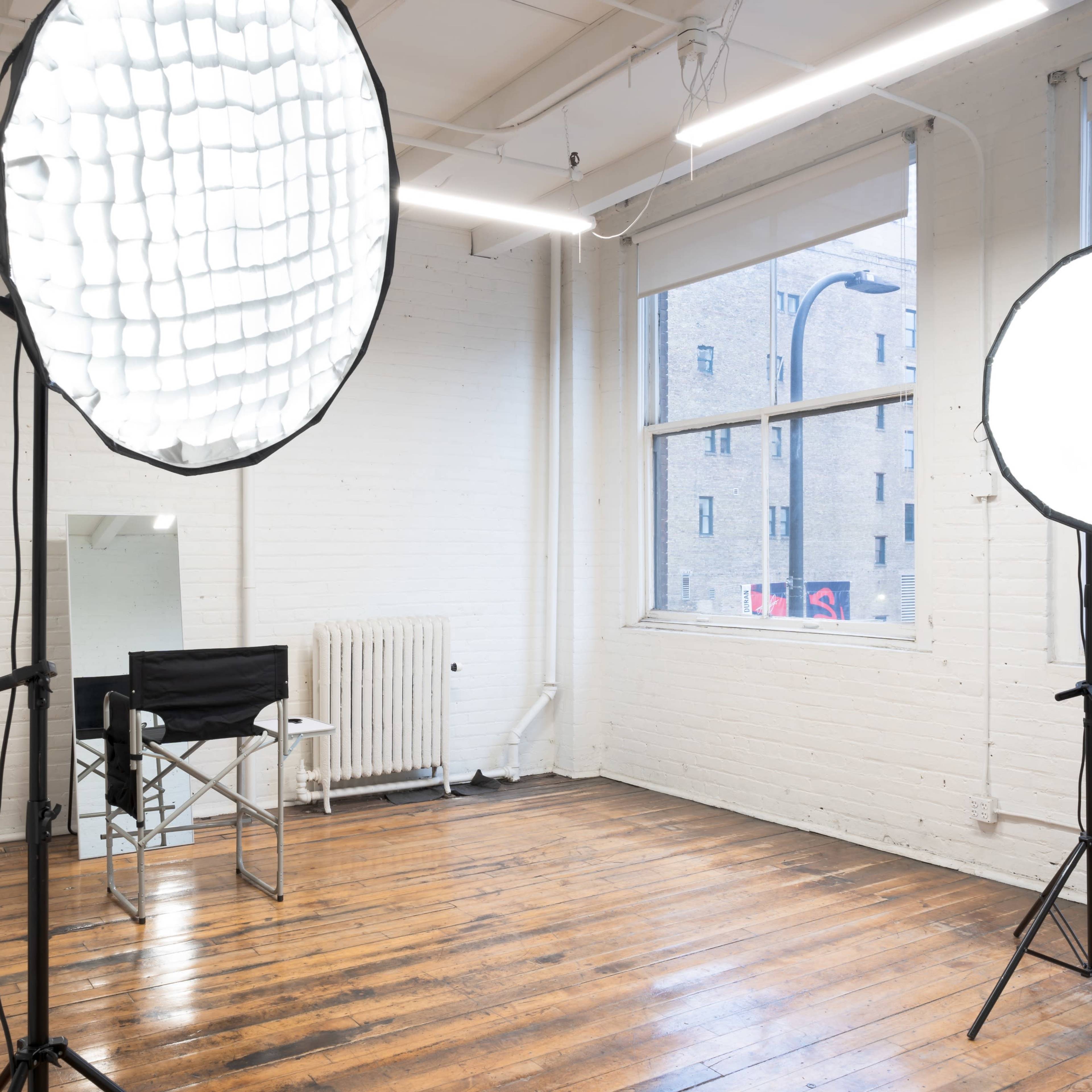A well-lit studio space with two large light softboxes, a black director's chair, a small table with a plant, and large windows revealing a city view.