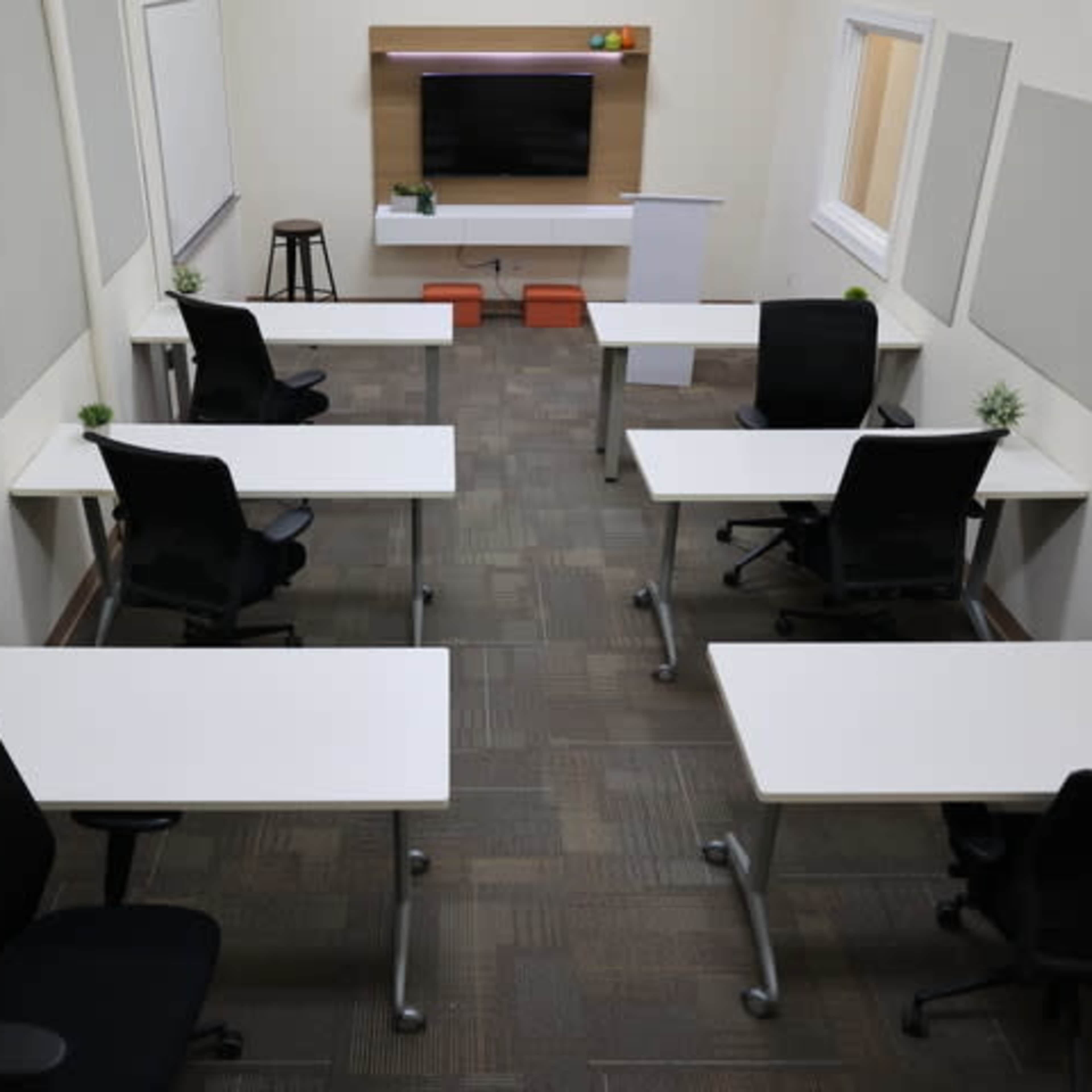A modern classroom or meeting room featuring several desks with chairs arranged around a central television and presentation area.