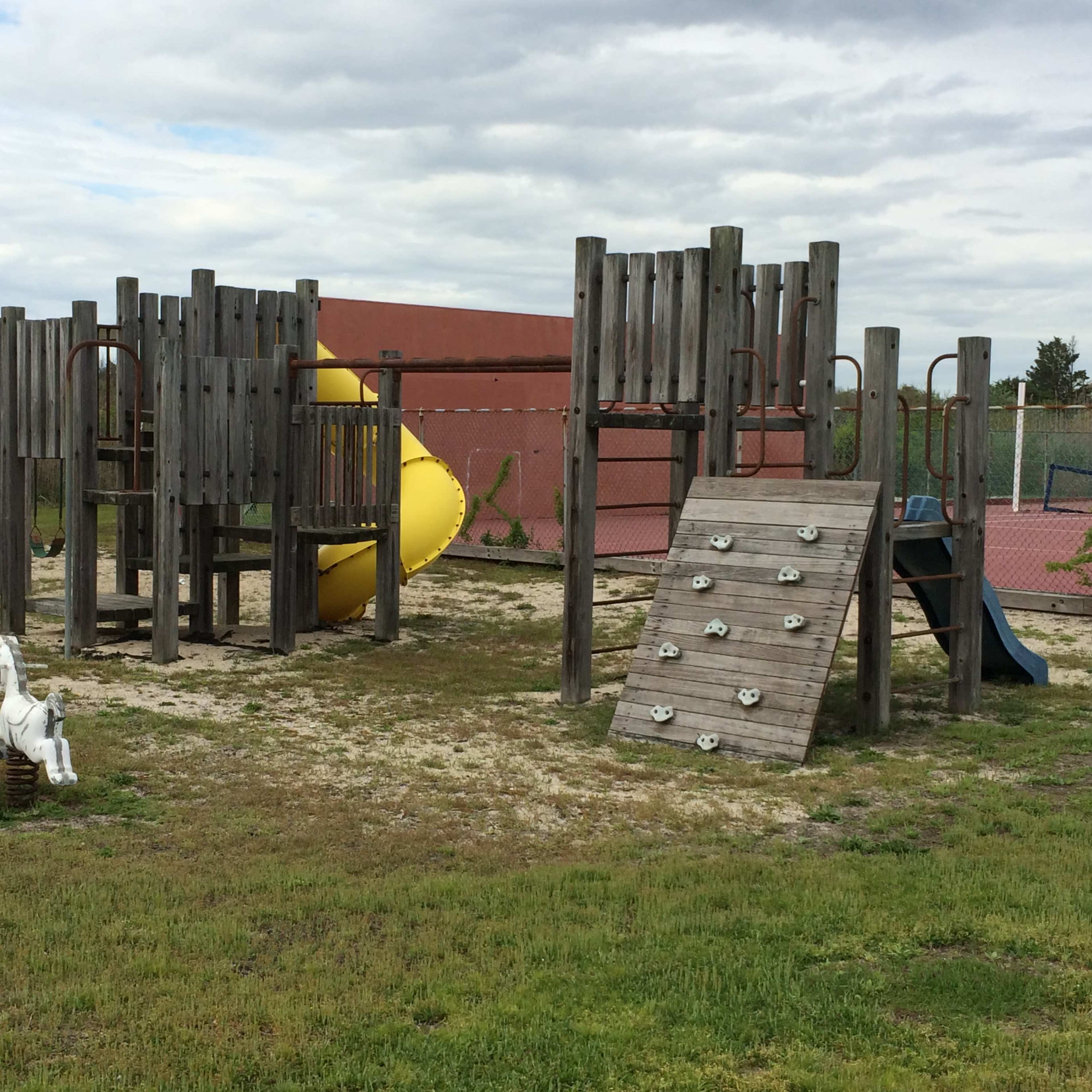 A playground with a slide, climbing wall, and a horse spring rider, set on sandy ground with a tennis court in the background.