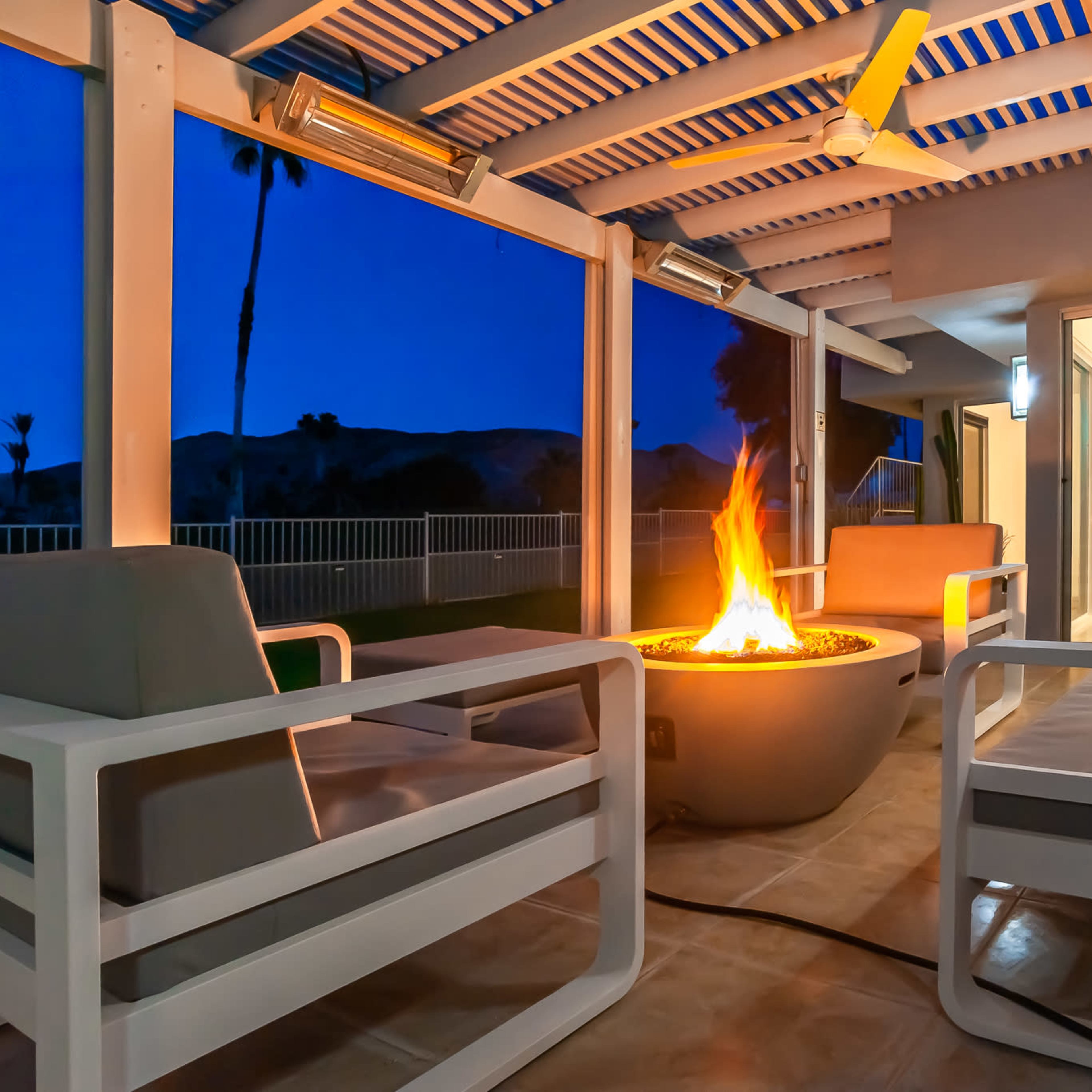 A cozy outdoor seating area features a circular fire pit surrounded by modern white chairs, under a pergola at dusk.