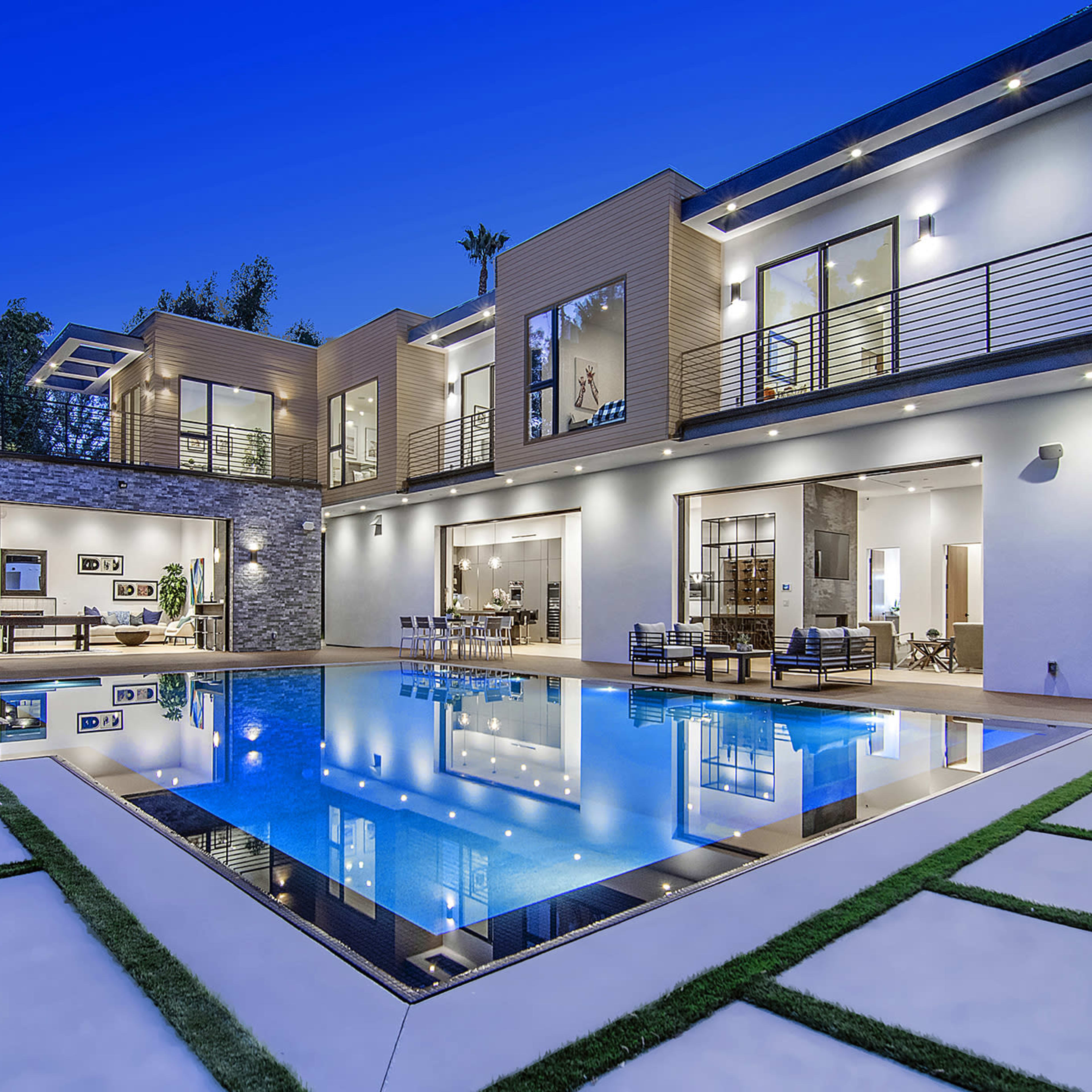 A modern house with large windows and a spacious pool area is illuminated at dusk, reflecting the surrounding landscape and architecture.