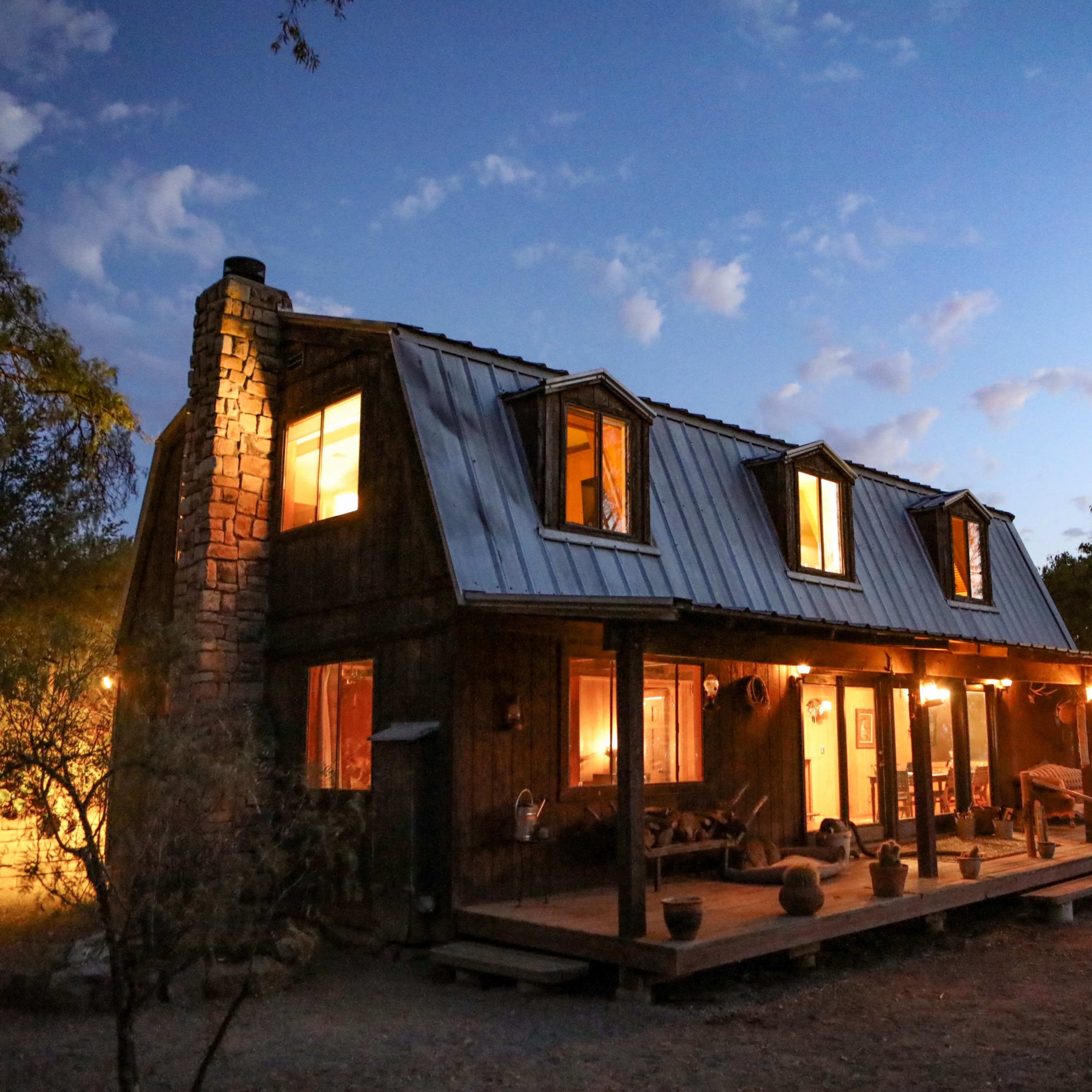 A rustic cabin with a metal roof and illuminated windows stands among trees at dusk.