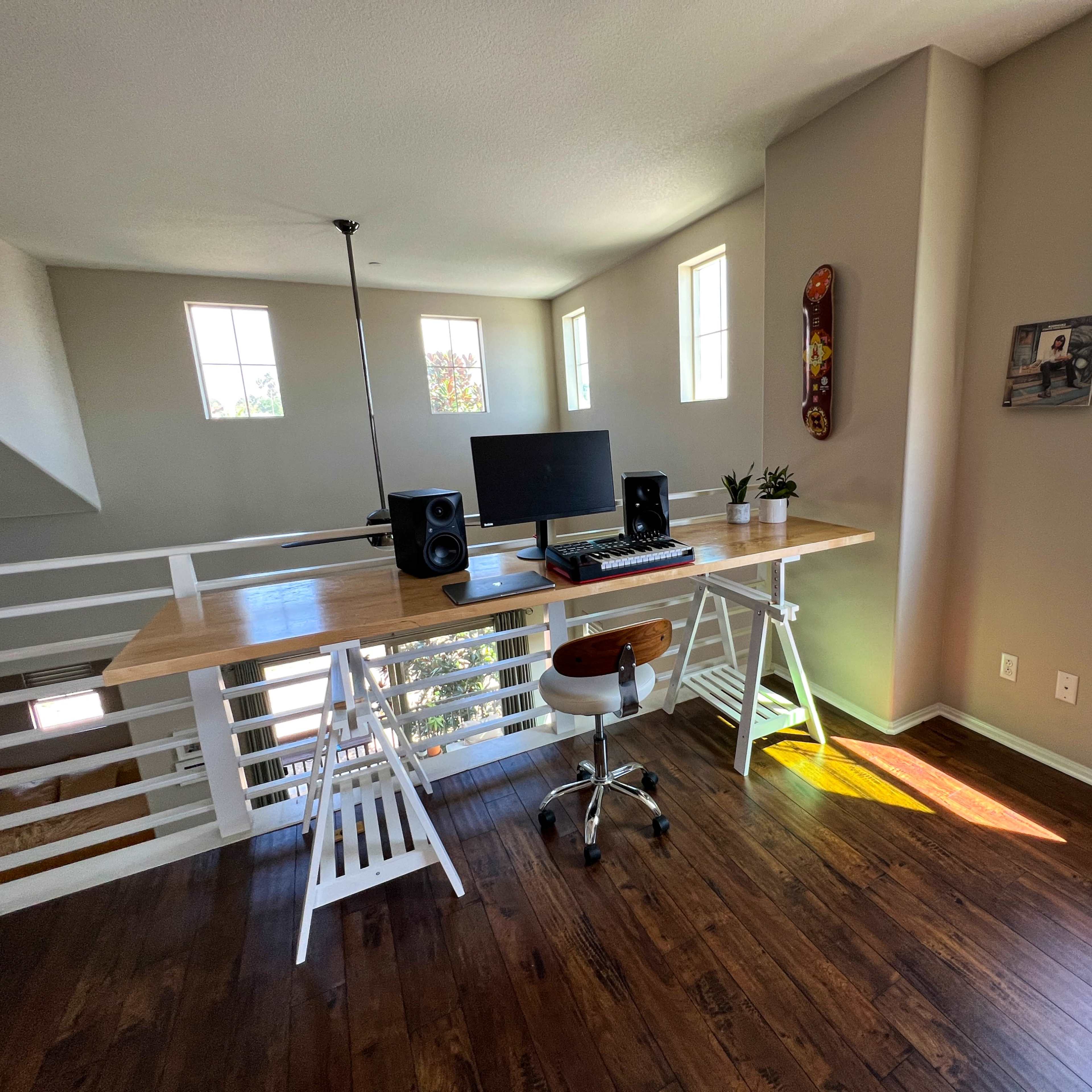 A minimalistic home office features a wooden desk with a computer, audio speakers, and a chair, situated in a brightly lit room with large windows.