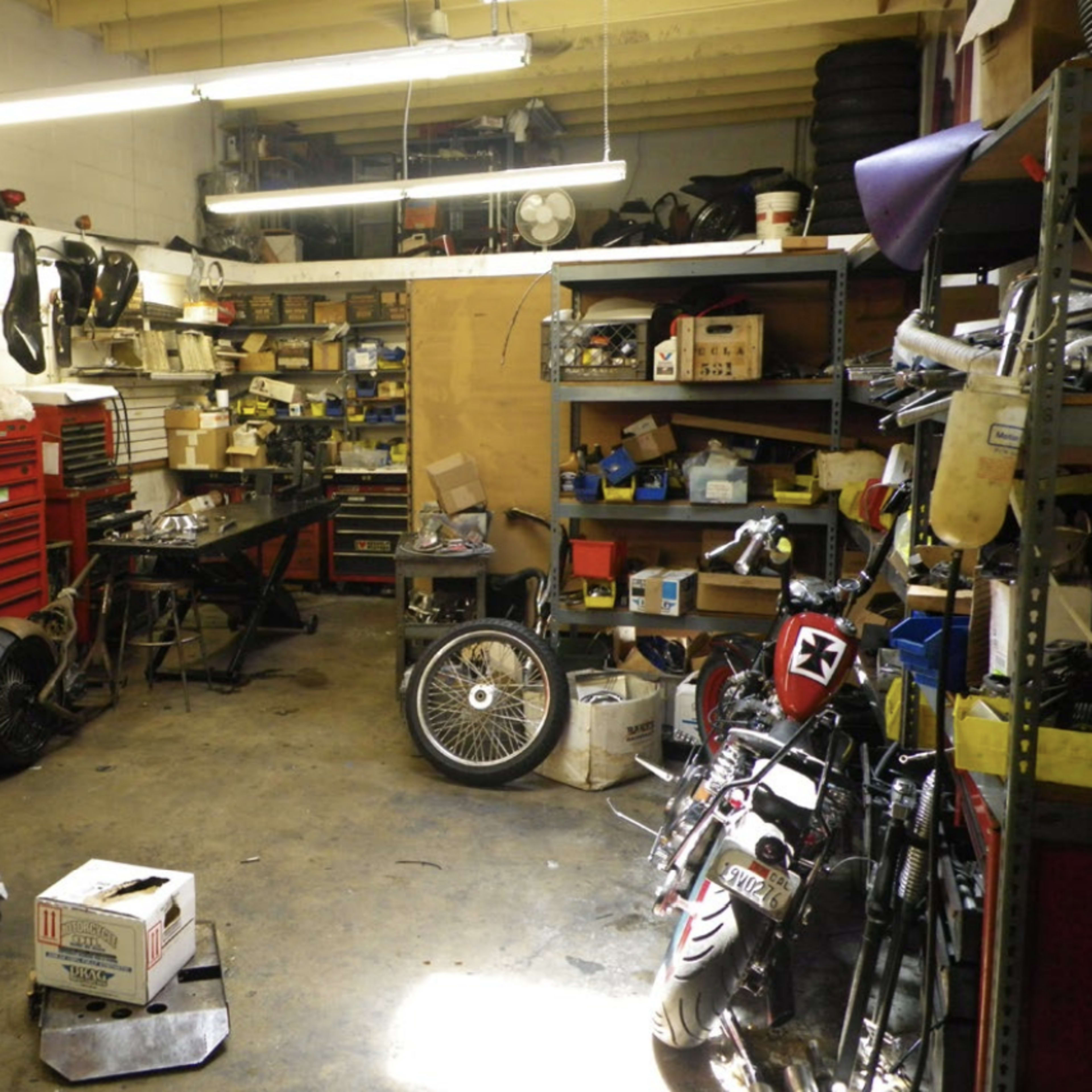 A cluttered garage workspace with various motorcycle parts, tools, and shelves filled with equipment.