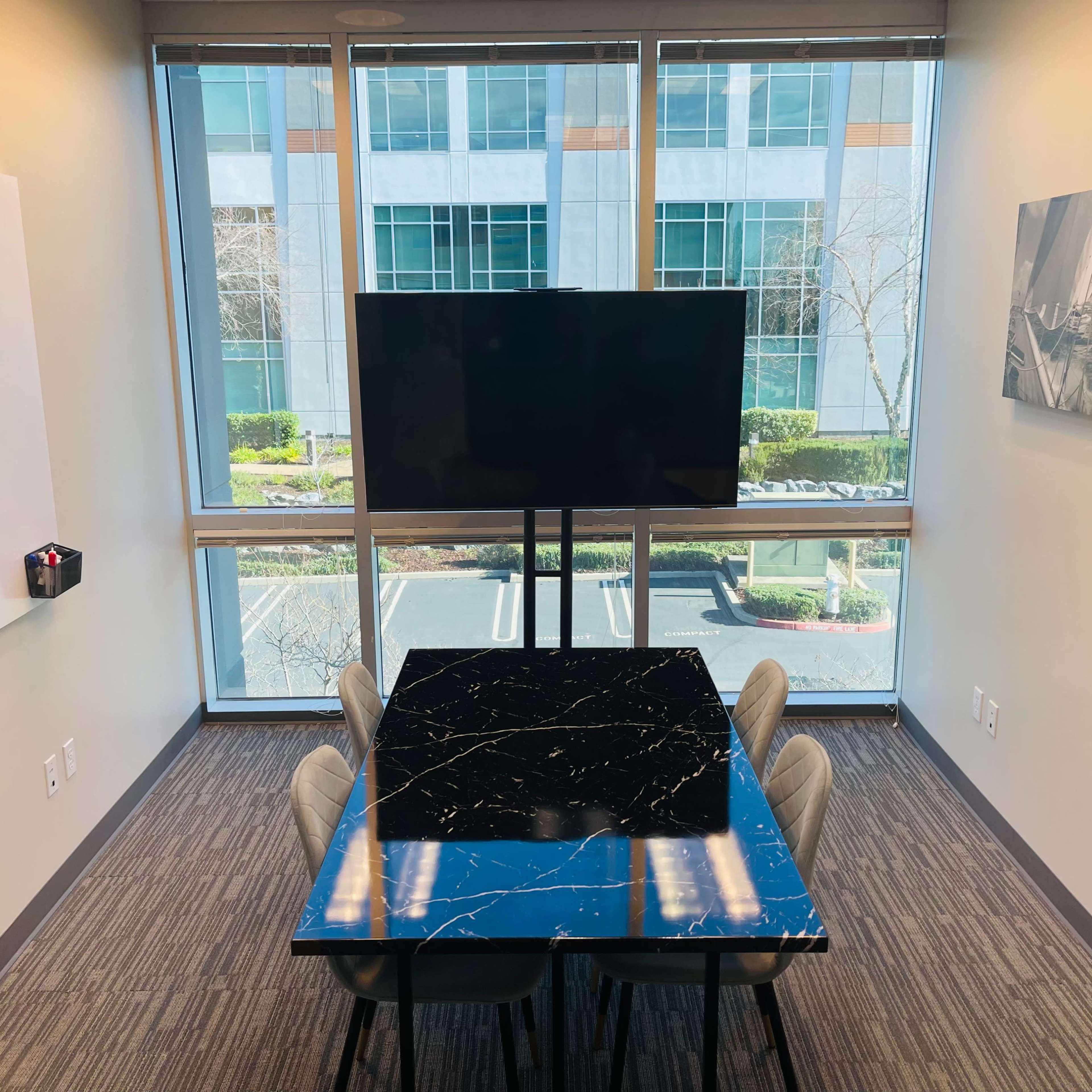 A modern conference room features a black marble table surrounded by light-colored chairs, a large TV on a stand, and large windows providing a view of the outdoors.