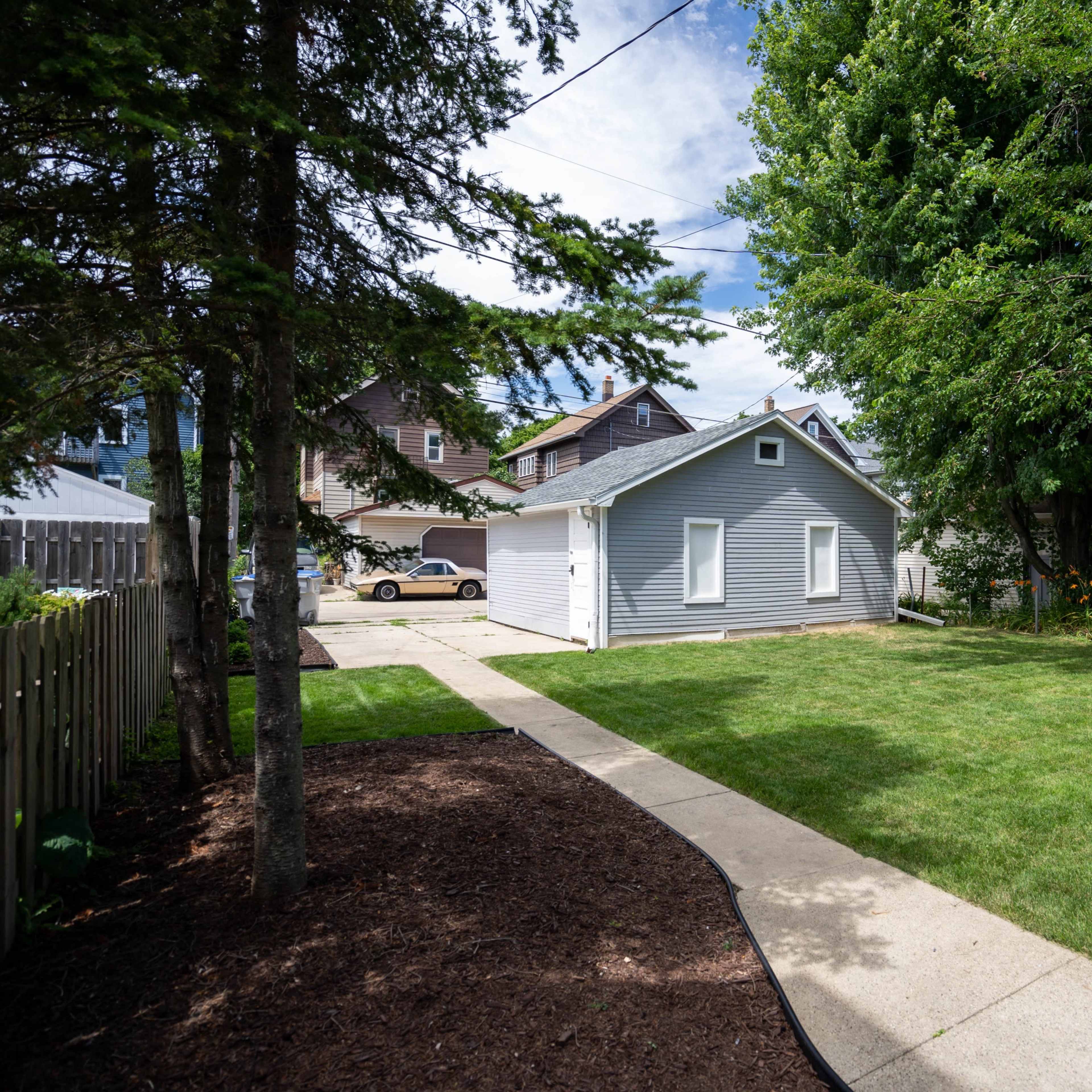 A landscaped yard with a concrete walkway leading to a gray shed, surrounded by trees and a few houses in the background.