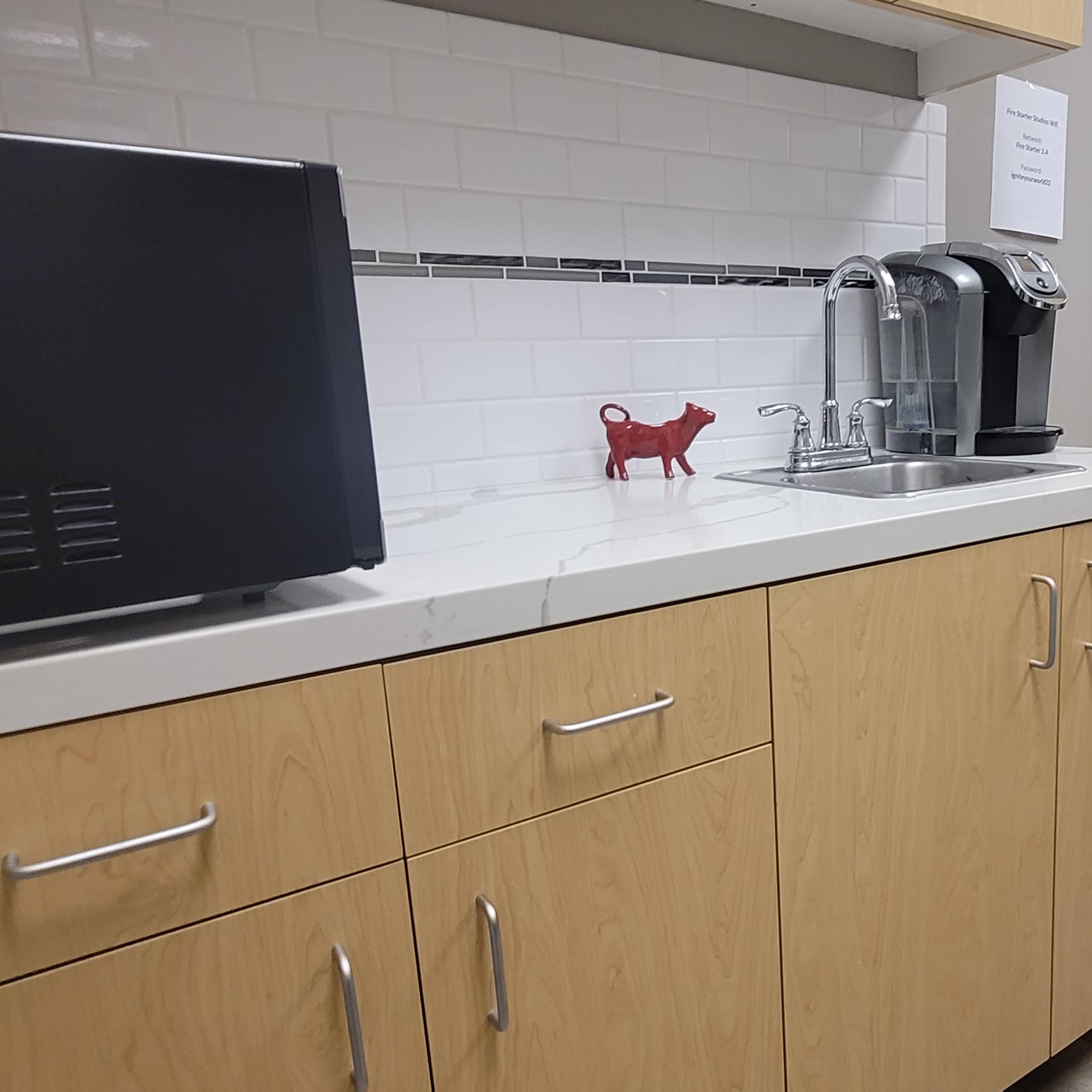 A kitchenette area with a black appliance, a red decorative item, and a coffee maker on a countertop, adjacent to a door leading to a hallway.