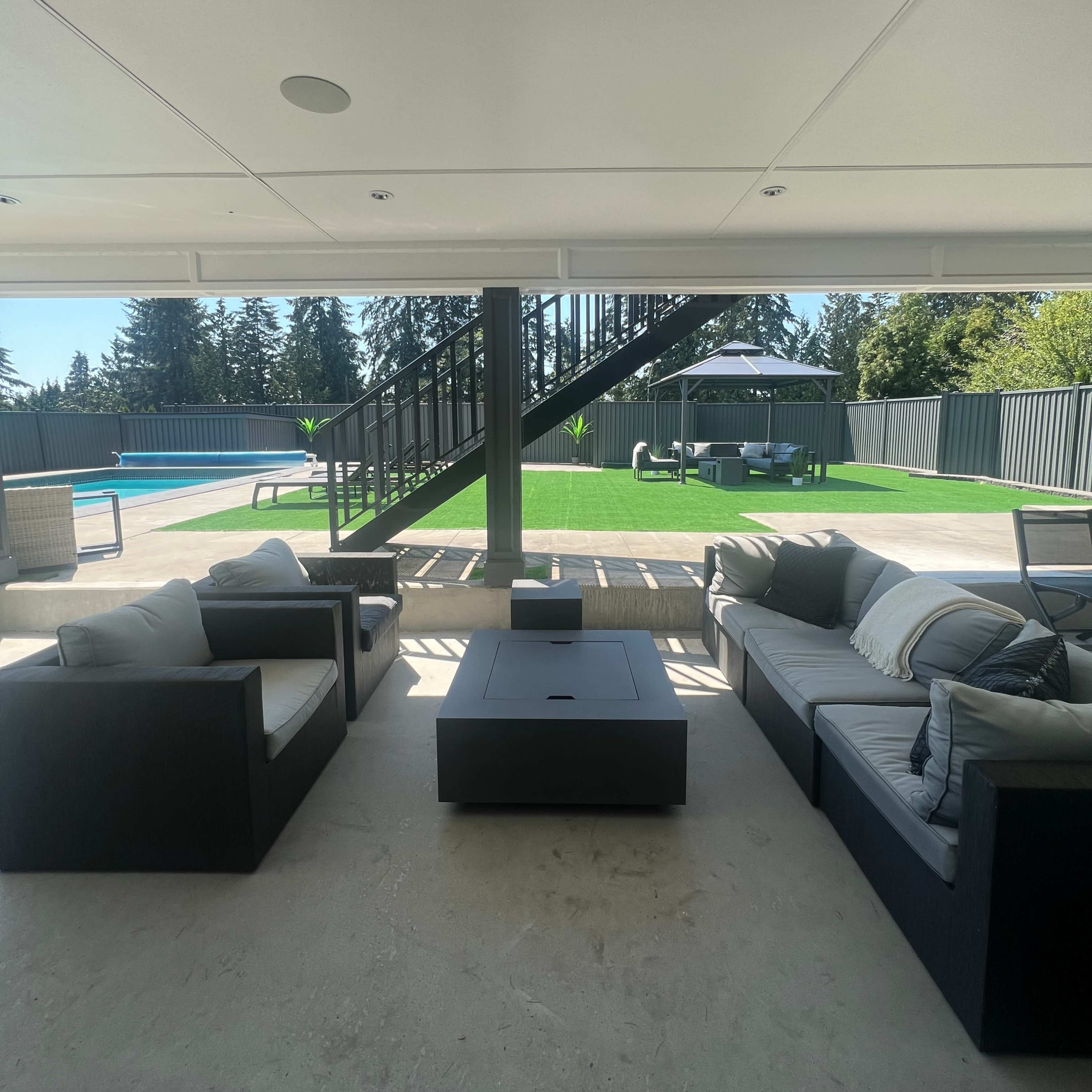 A modern outdoor living area with black furniture facing a pool and a grassy yard, framed by a staircase in the background.