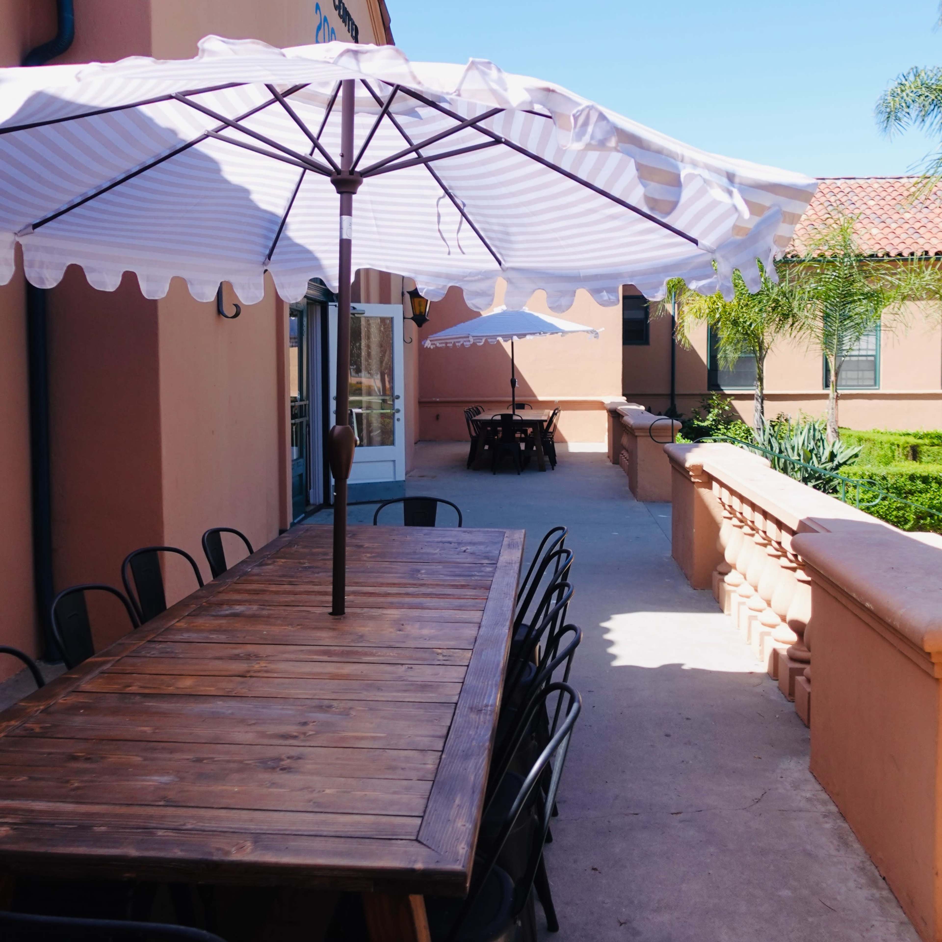 A wooden table with black chairs and large umbrellas is situated along a pathway next to a building with a stucco exterior.