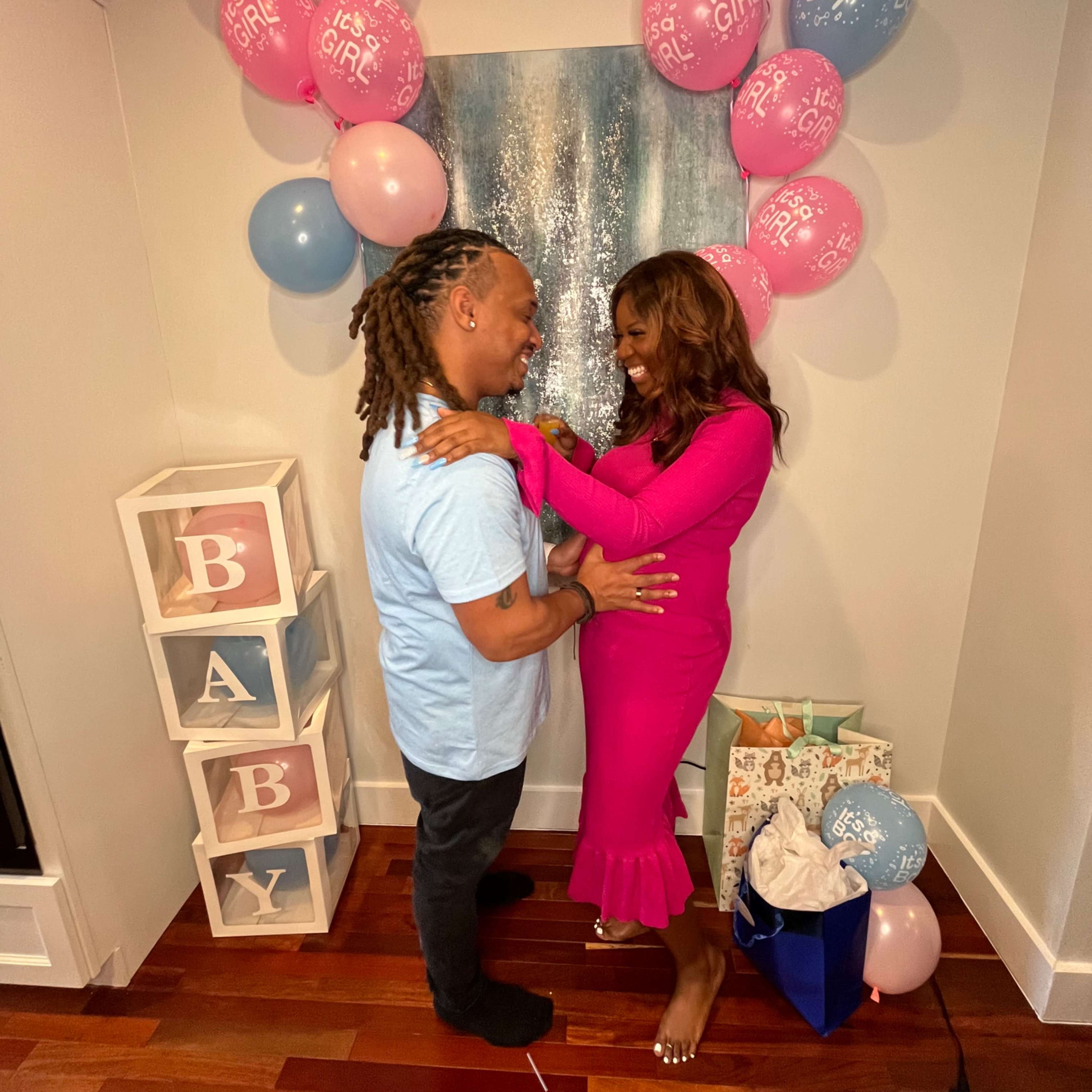 A couple stands together in a decorated room with pink and blue balloons and a display that reads "BABY."
