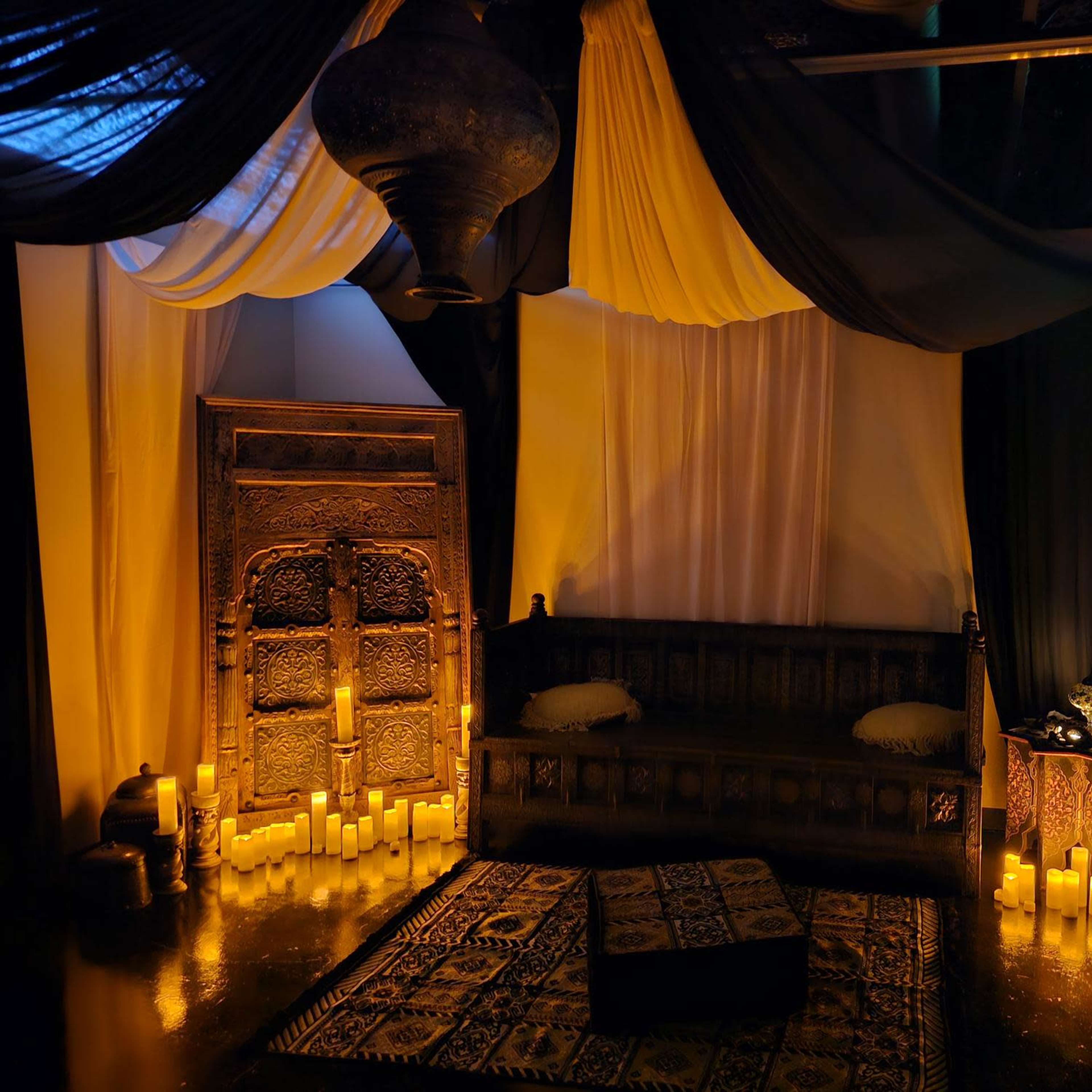 A softly lit interior with a decorative wooden couch, surrounded by candles and draped fabric in various colors.