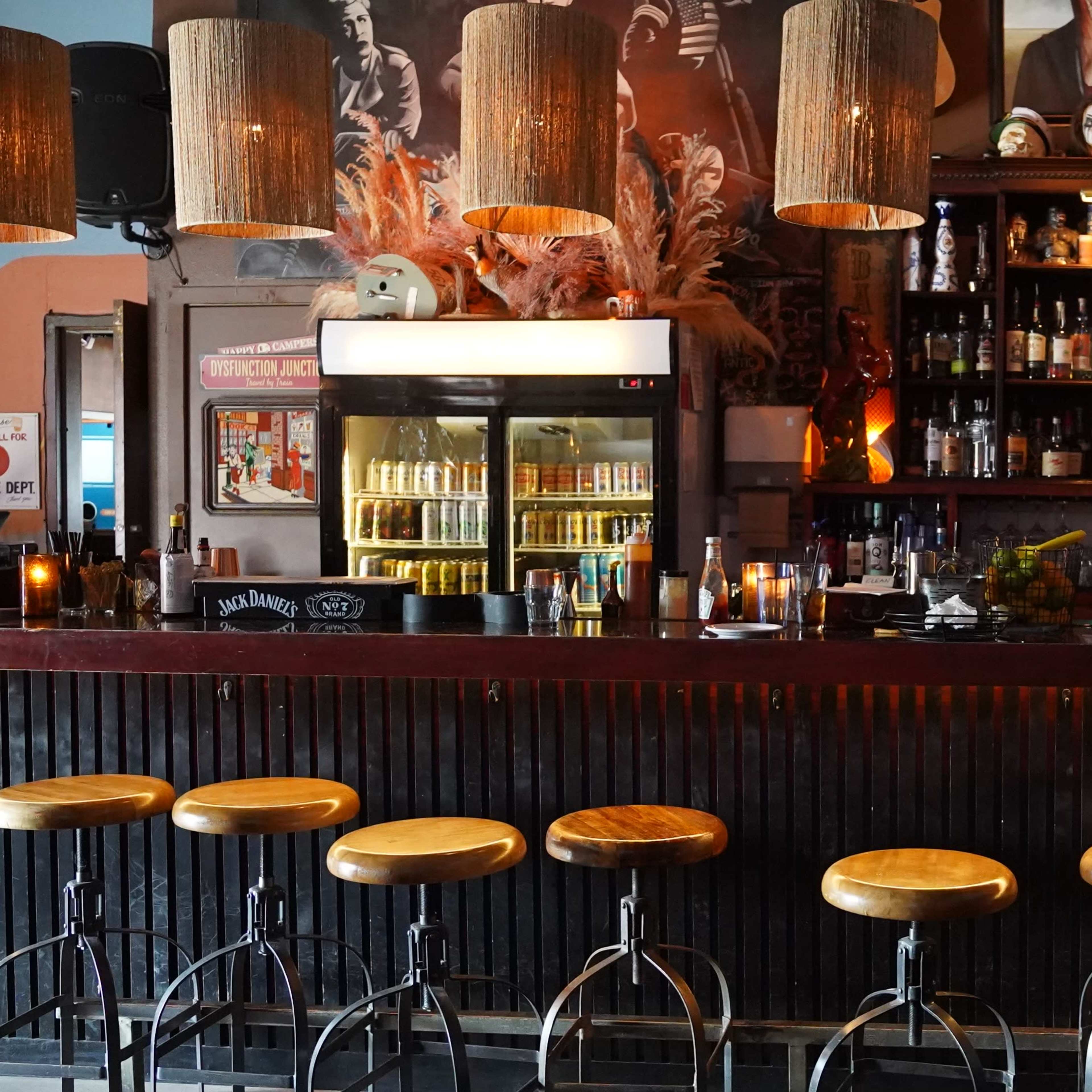 A bar with wooden stools lined up in front of a dark wooden counter, illuminated by pendant lights and featuring shelves filled with various bottles behind it.