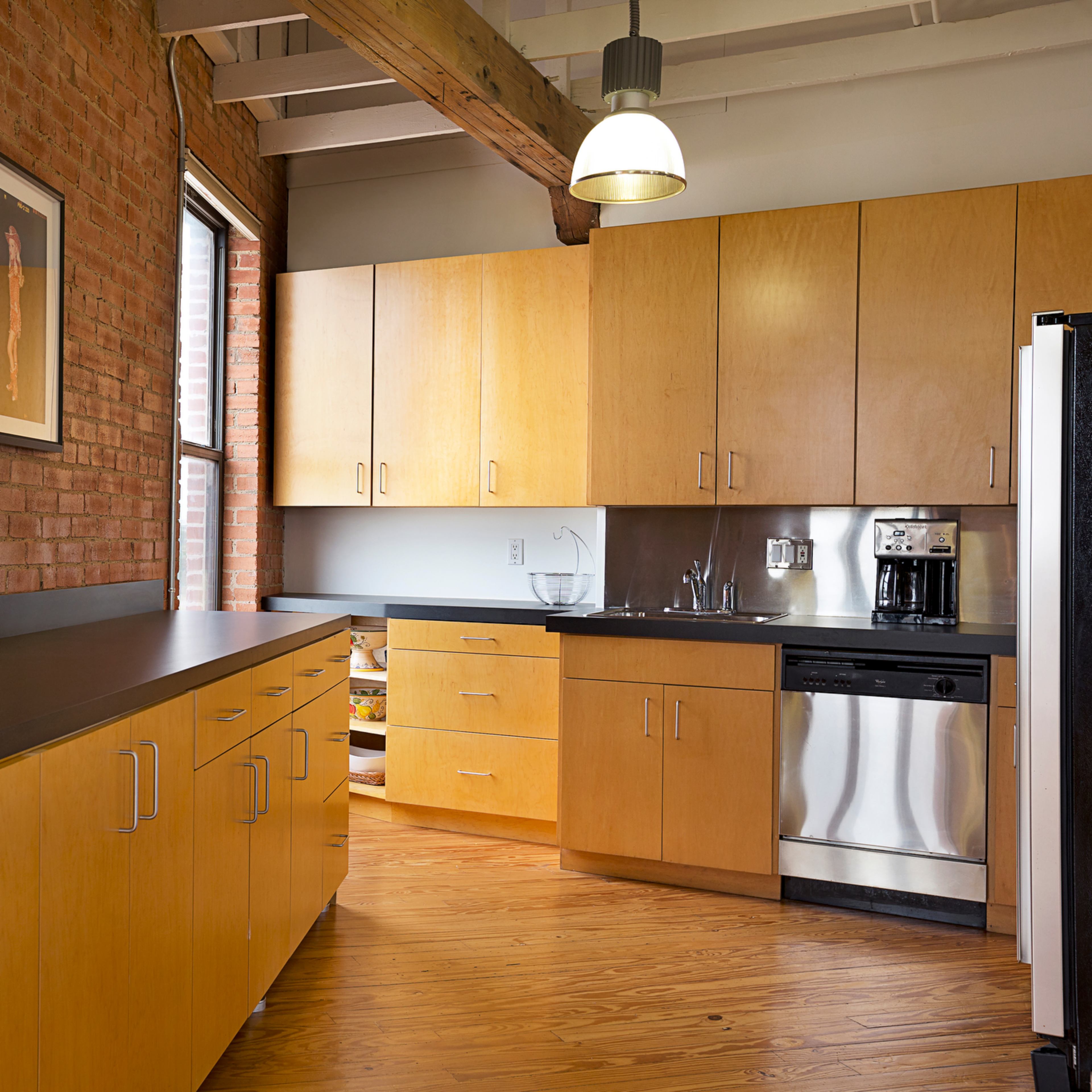 A modern kitchen featuring wooden cabinetry, a stainless steel refrigerator, and a large window with exposed brick walls.
