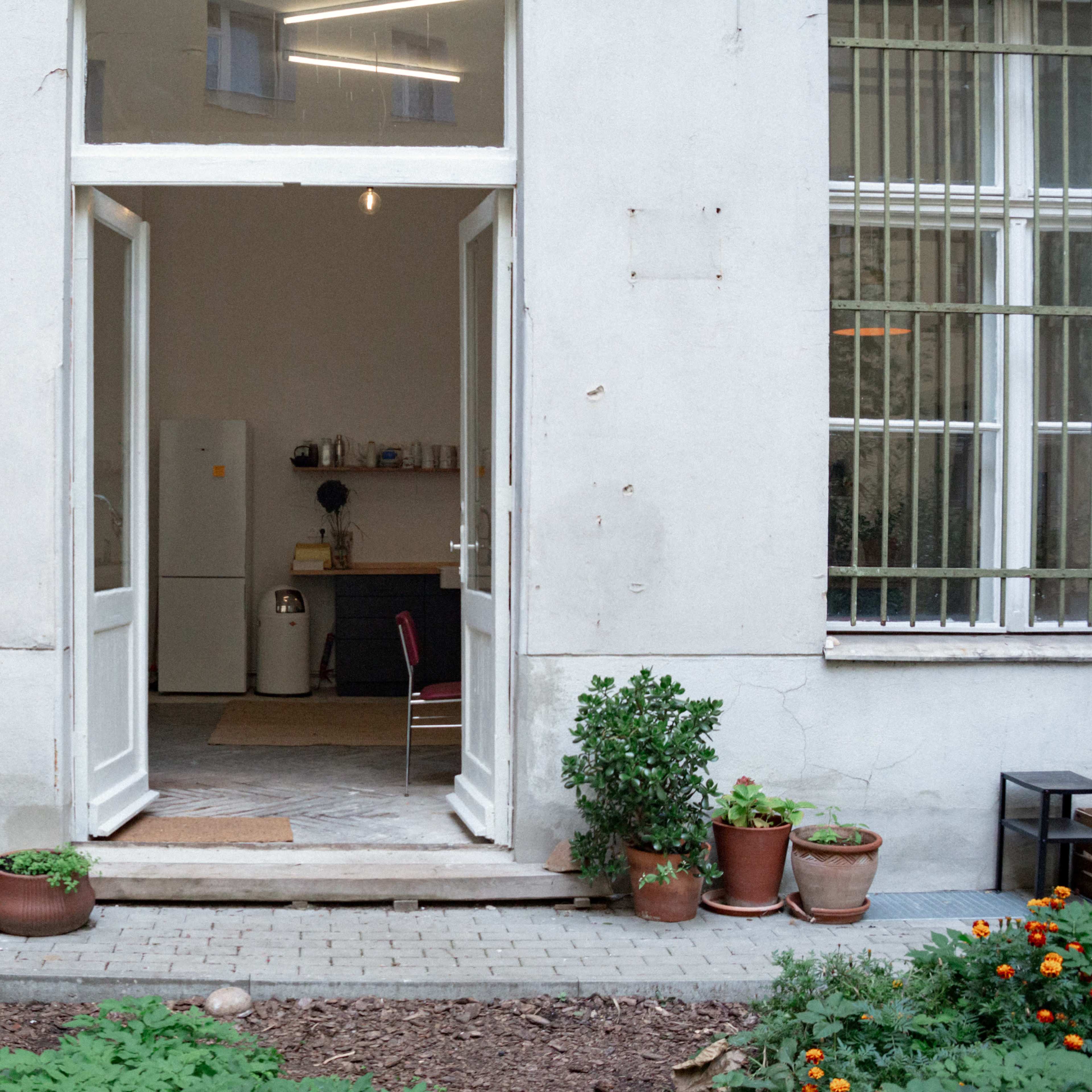 A bright, open doorway leads into a spacious kitchen area with white and neutral furnishings, surrounded by potted plants on a stone pathway.
